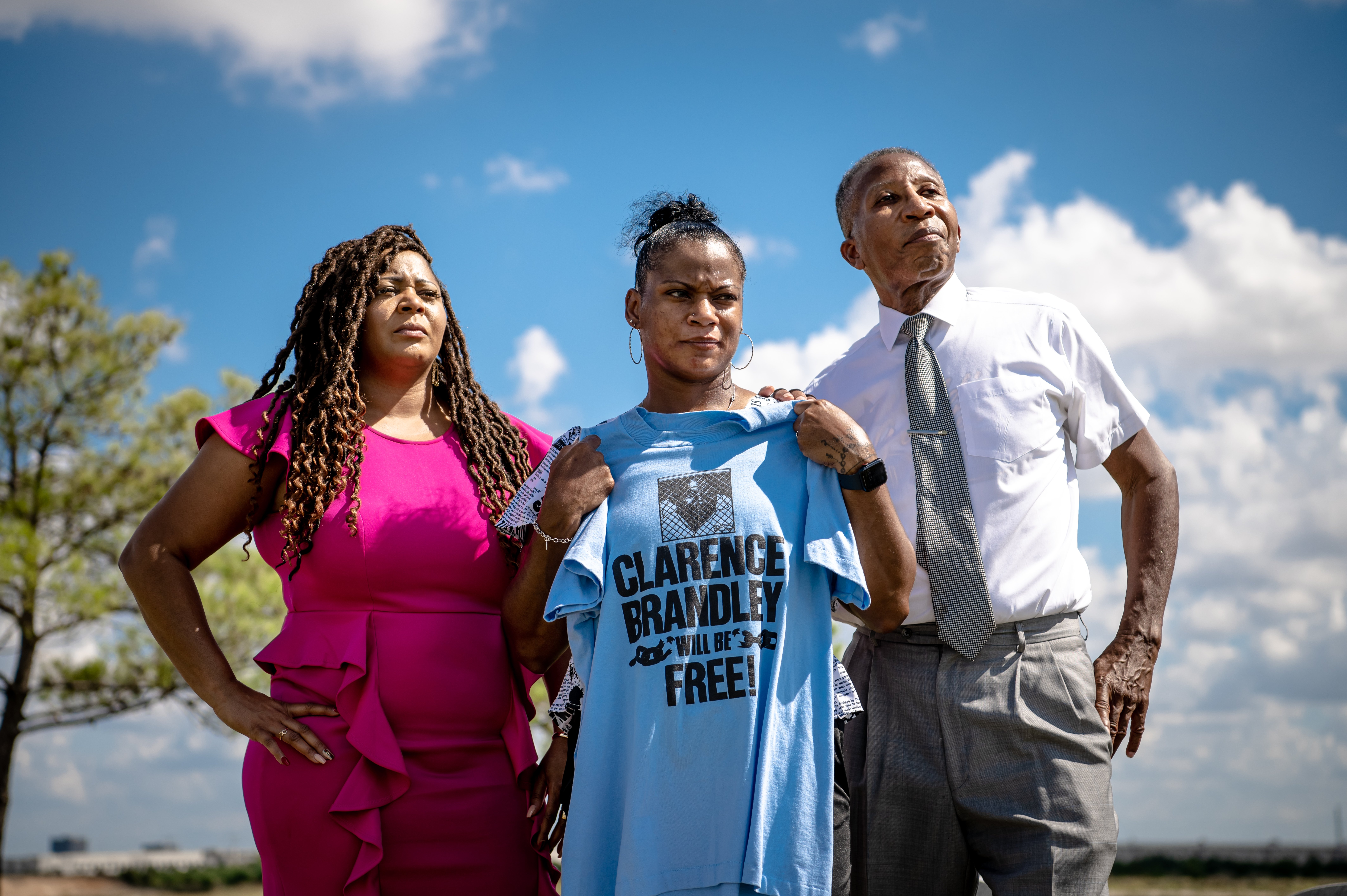 Ozell Brandley (a Black man) and his nieces, Cassie Brandley and Elizabeth Phillips, visit the grave of Clarence Brandley. Elizabeth Phillips is holding a blue t-shirt which reads, "Clarence Brandley Will be Free."