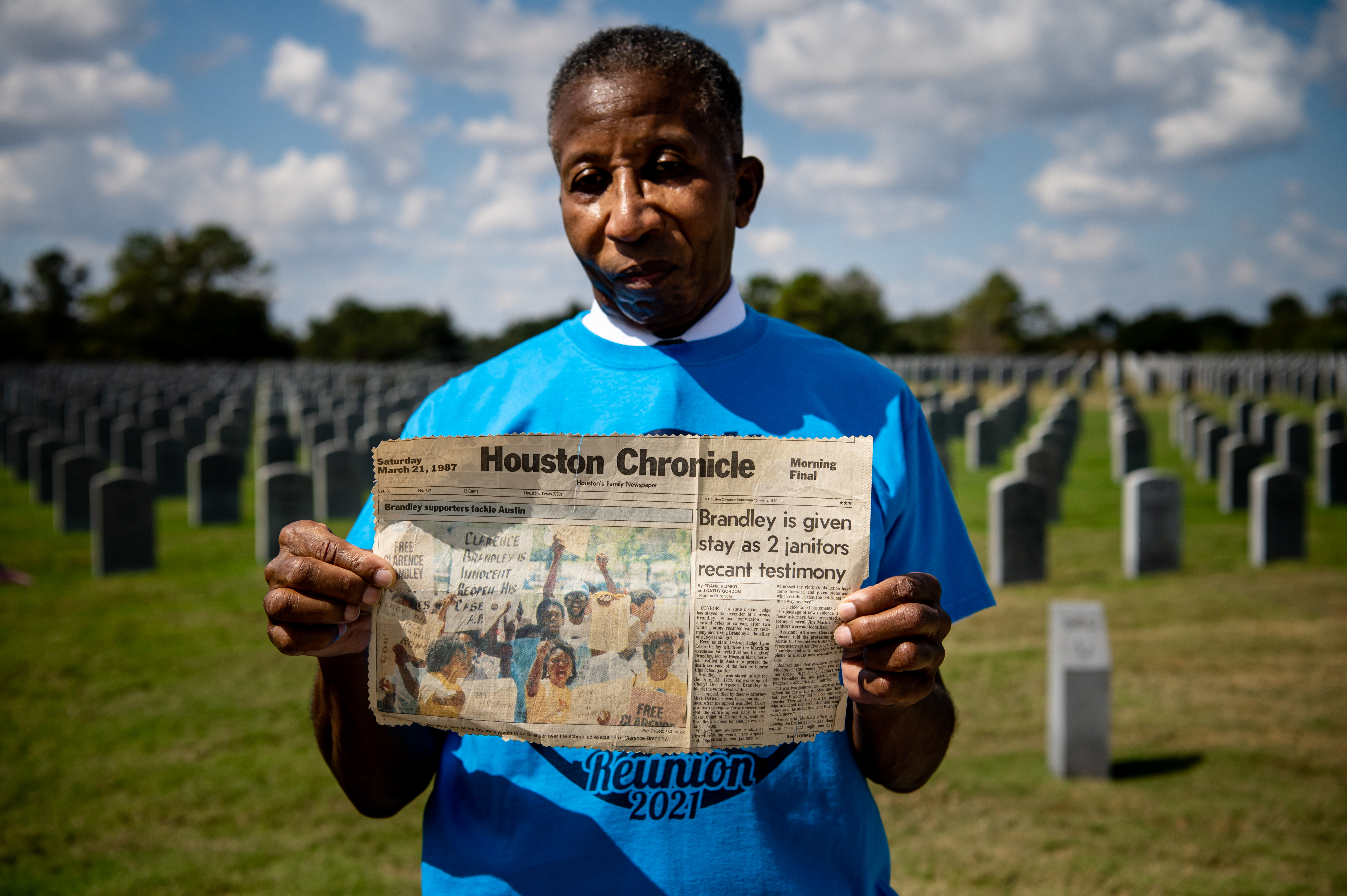 An older Black man, standing in a sunlit cemetery, holds up a newspaper clipping which reads "Brandley is given stay as 2 janitors recant testimony."