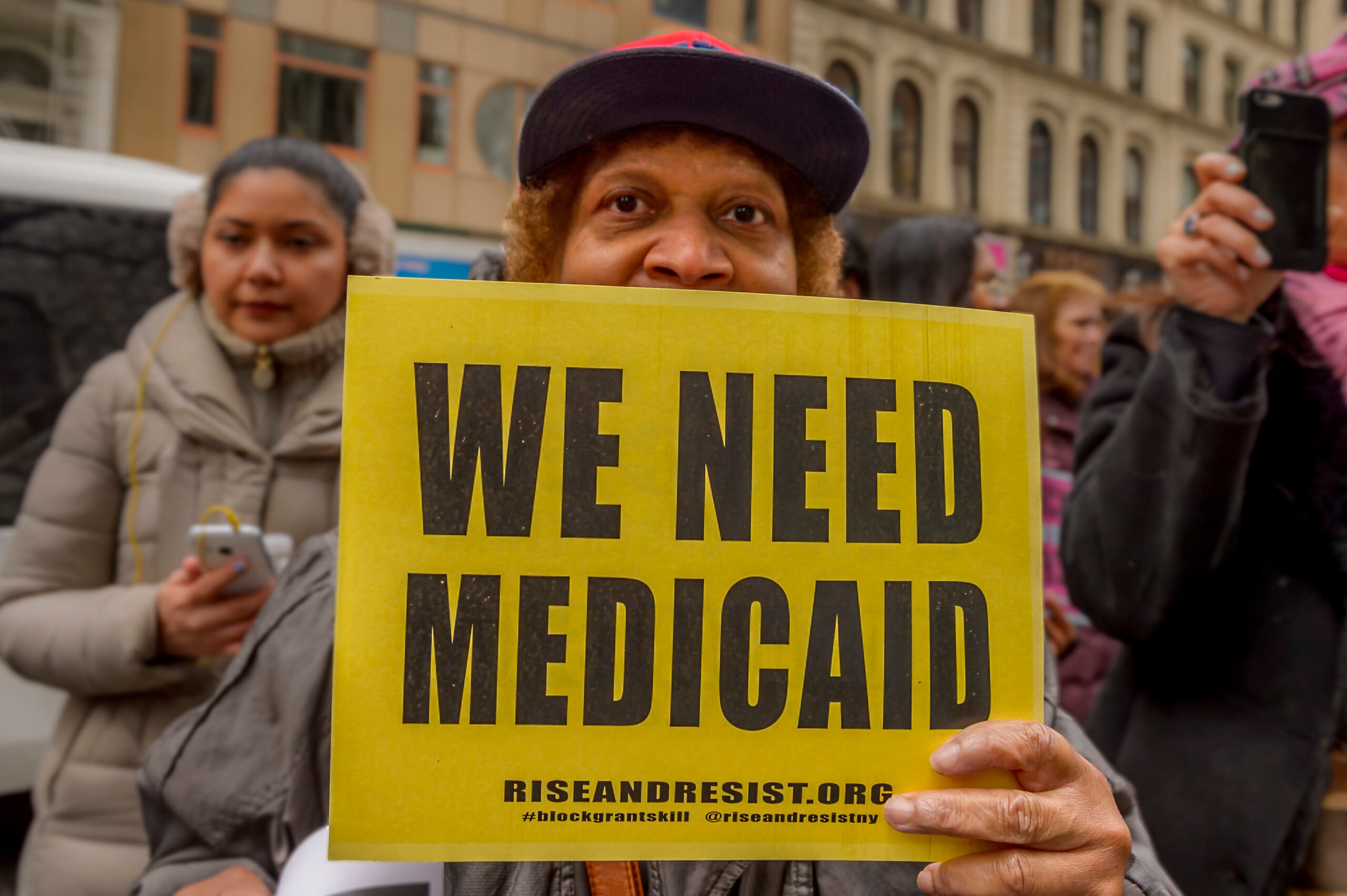 A BIPOC activist holds up a yellow sign which reads "We Need Medicaid" in a tightly framed photo of a protest.