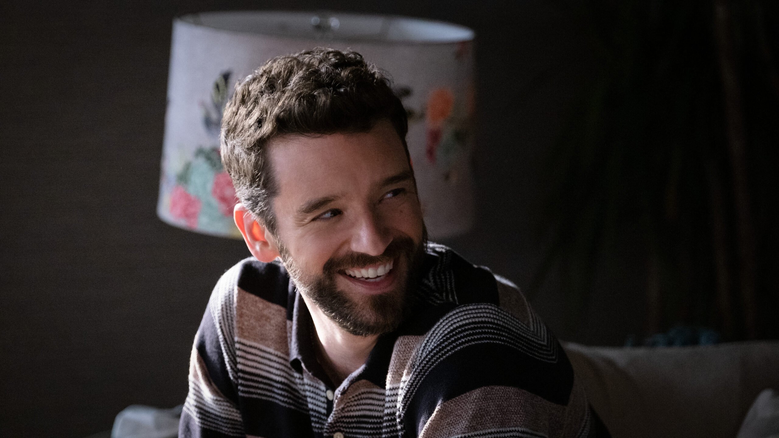 Michael Urie in an appearance in Apple TV's Shrinking, a smiling white man with full beard and mustache, wearing a blue and gray striped polo shirt.