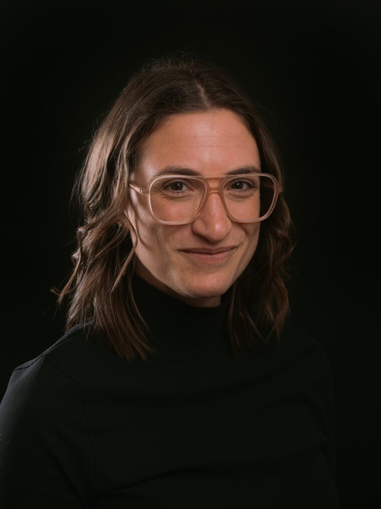Nikki Kobiljak has shoulder length brown hair, large clear plastic glasses, and a slight smile on her face. She's wearing a dark turtleneck.
