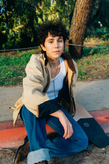 Olive Klug sits outside on a curb, wearing blue jeans with rolled cuffs, a tan jacket, and a scoop neck white tee. They are a nonbinary white person with curly dark brown hair.