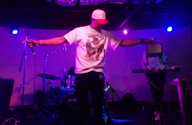 David Shabani holds his arms extended, looking downward and away from the camera, the microphone in one hand. He's a Black man wearing a vintage Michael Jackson tee, a baseball cap and shades.