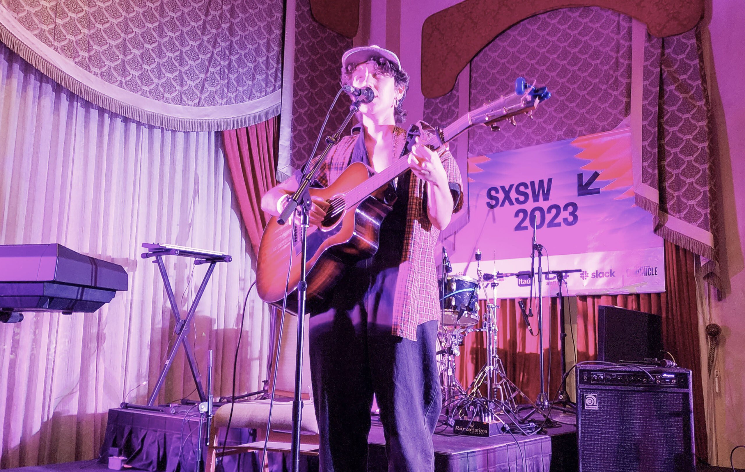 Olive Klug is a white nonbinary person wearing a floppy newsie hat over their curly dark brown hair, a flannel shirt, a v-neck tee and baggy blue jeans. They are playing a guitar on stage alone, surrounded by instruments. On the walls are antique looking draperies and a SXSW 2023 banner.