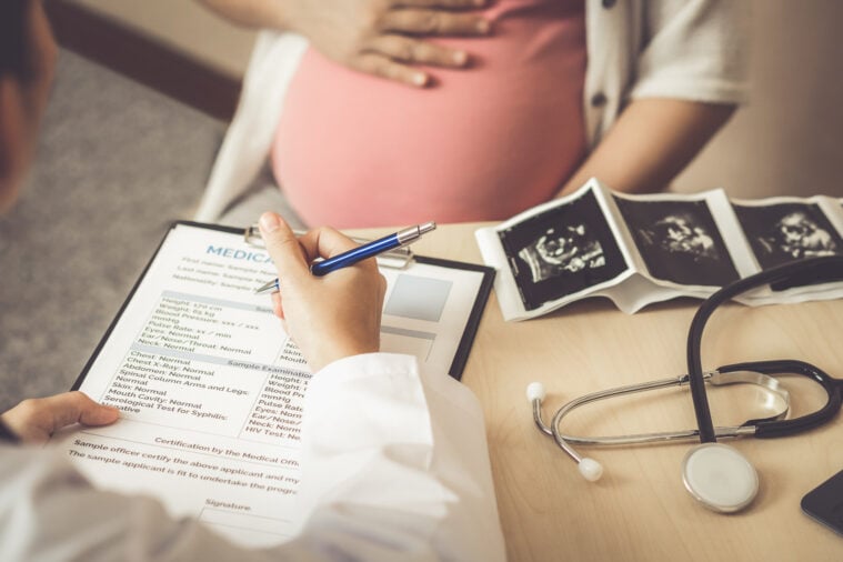 Stock image of a pregnant person in a doctor's office, with a doctor holding a clip board of medication instructions. There are sonogram results on a table, along with a stethoscope.