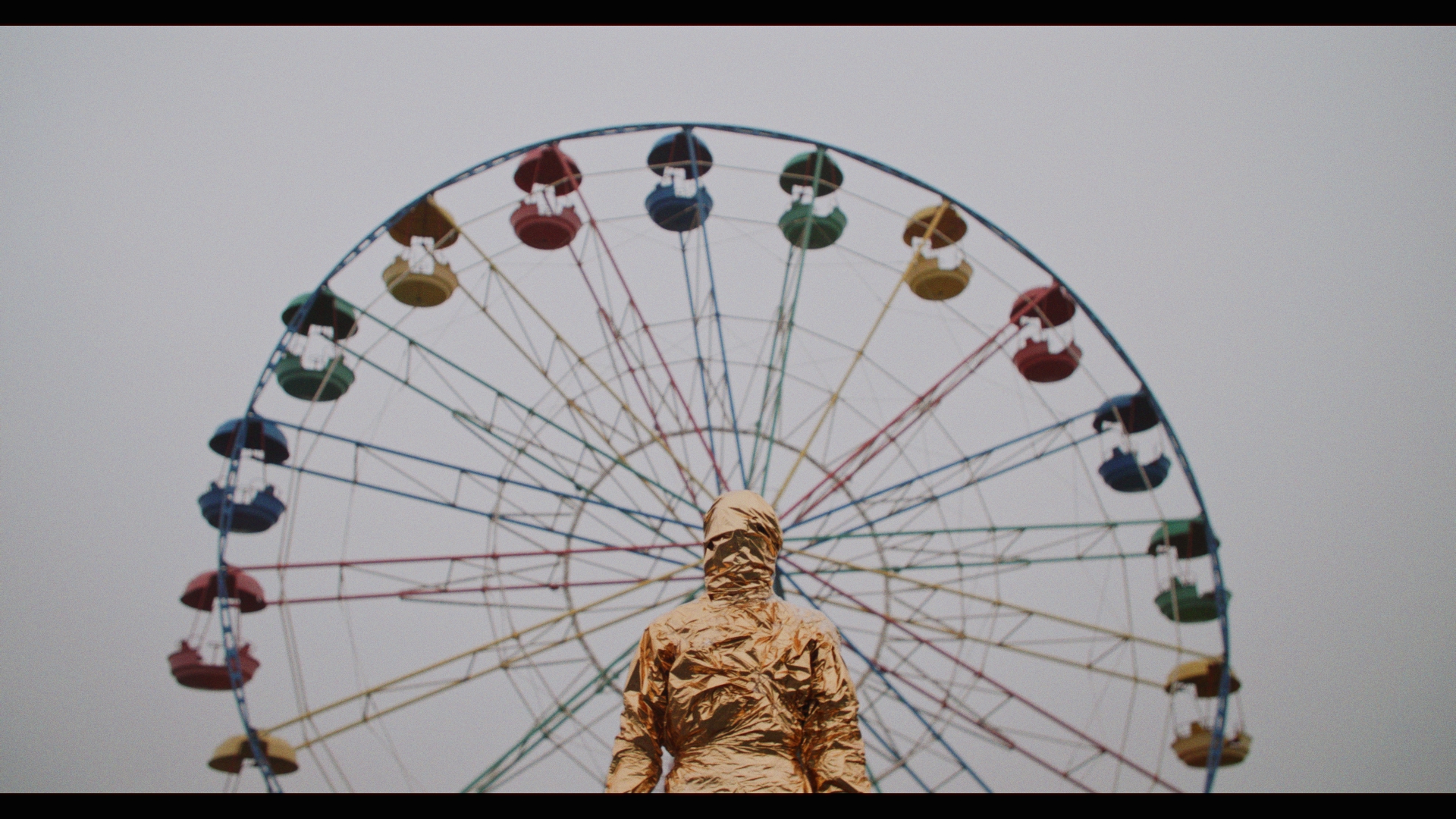 Performer Gena is standing in front of a ferris wheel, looking mummified with her body completely covered in off-white wrappings. The sky is a featureless gray.