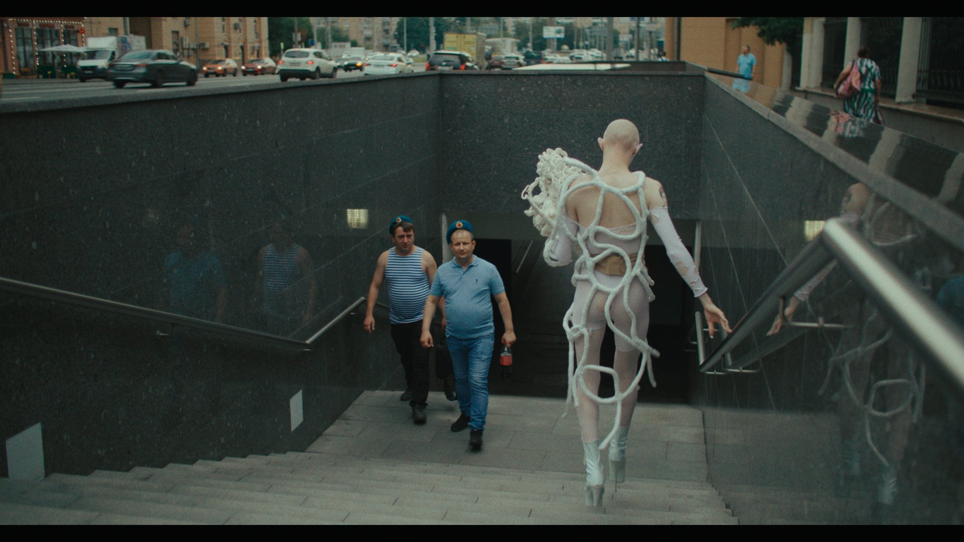 Gena, seen from behind, is wearing a torn body stocking under a complex, root-like white costume. She is bald and carrying her custom wig in her hands. She's wearing high-heeled boots and carefully walking down steps into Moscow's underground, as two onlookers stare.
