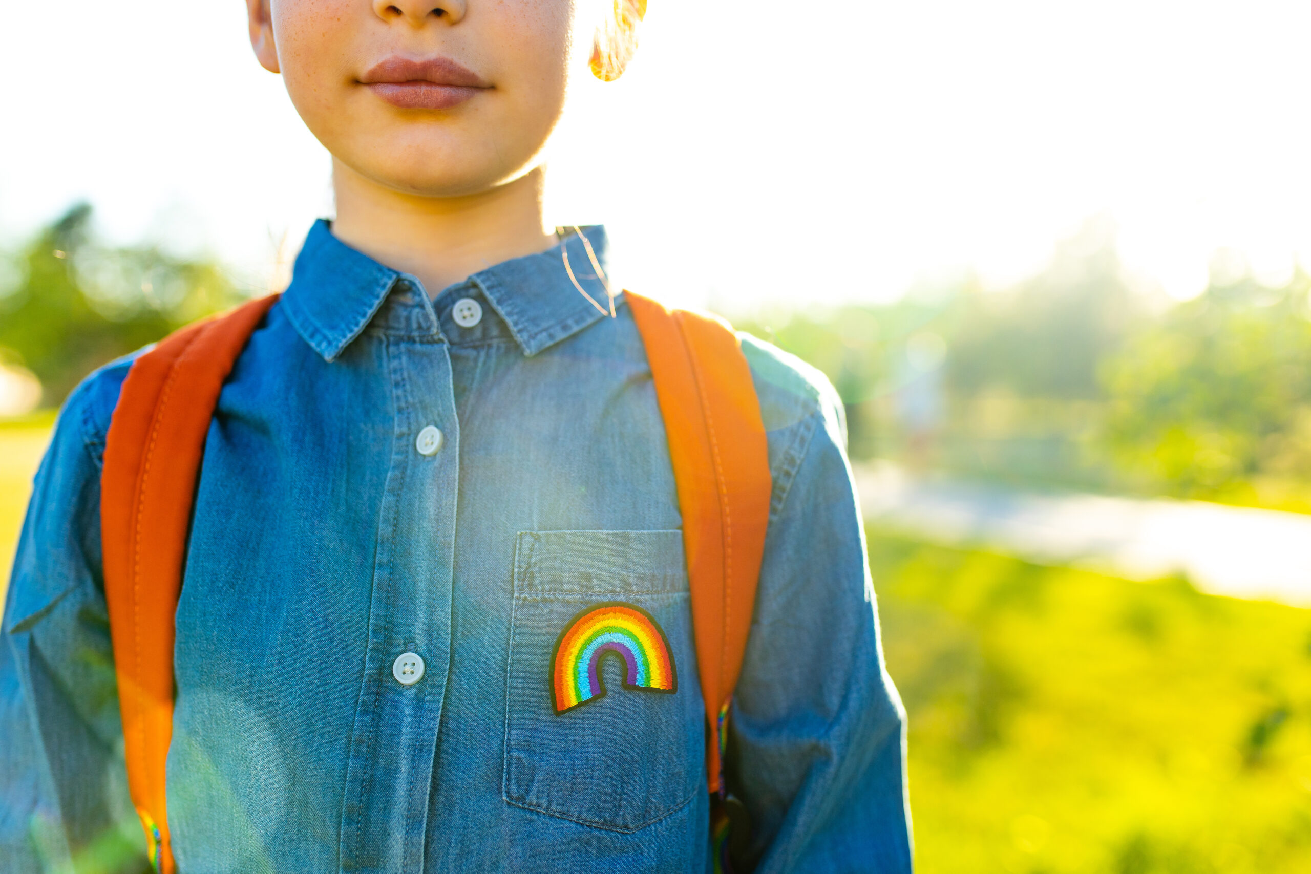A pre-adolescent child in a denim jacket with a rainbow Pride patch, with red backpack straps visible. Just their nose and mouth are visible at the top of the photo. Texas is making life unbearable for transgender and LGBTQ+ students.