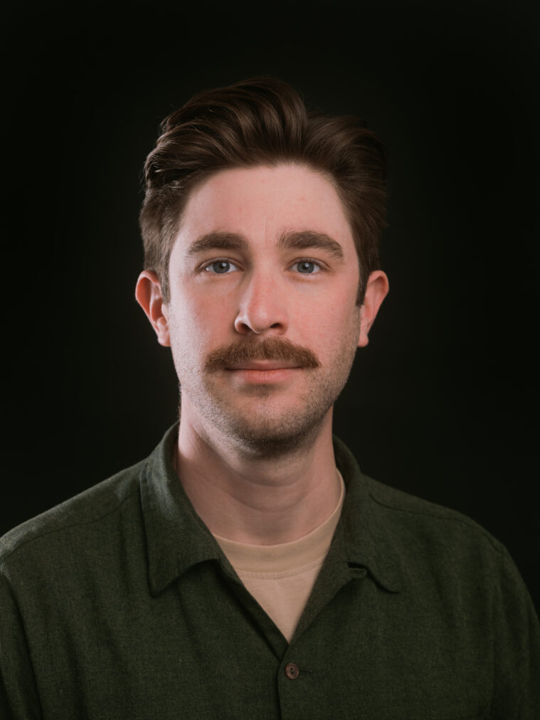 Steven Monacelli is a white man with short brown hair and a curved brown mustache. He's wearing a green v-neck.