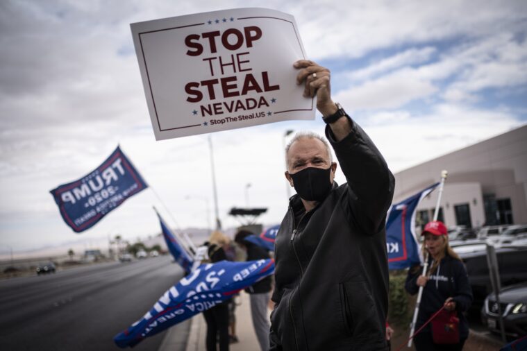 Trump supporters rally outside with flags for the president. In the foreground, one man in a mask holds a "Stop ted he Steal" sign.