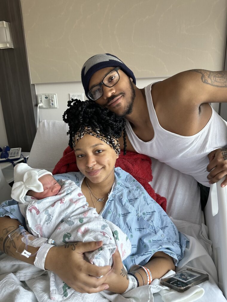 A Black couple pose proudly with their swaddled newborn baby.