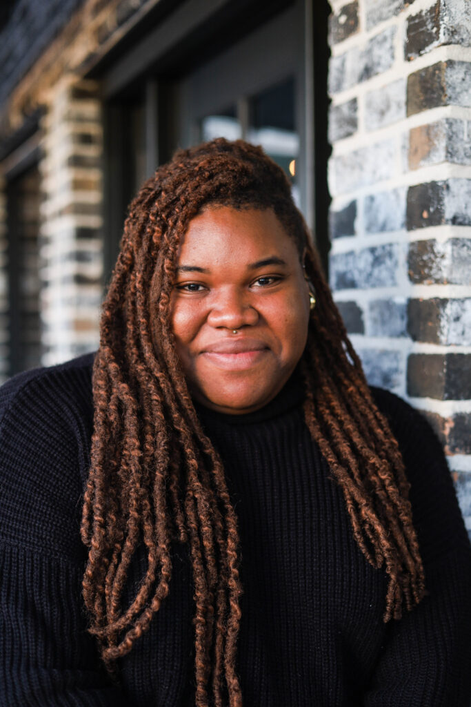 Tyler R. Lewis is a nonbinary Black person with braided hair and a silver stud medusa piercing. They are wearing a black top and standing in front of a brick house.