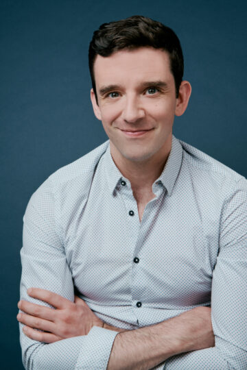 Michael Urie, a white man with short dark hair, has his arms crossed, wearing a polka dot button down shirt.