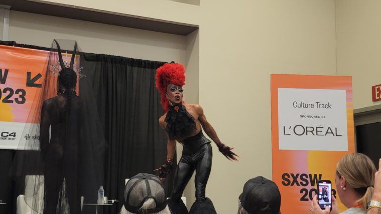 A Black drag performer in a flamboyant red wig, tight leather leather pants, and corsetted top performs on stage at SXSW. Another performer has her back to the audience.