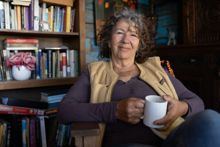 Diane Wilson, an older gray-haired woman in a study with a stuffed bookshelf behind her. She's sitting in an armchair holding a coffee cup and smiling slightly.