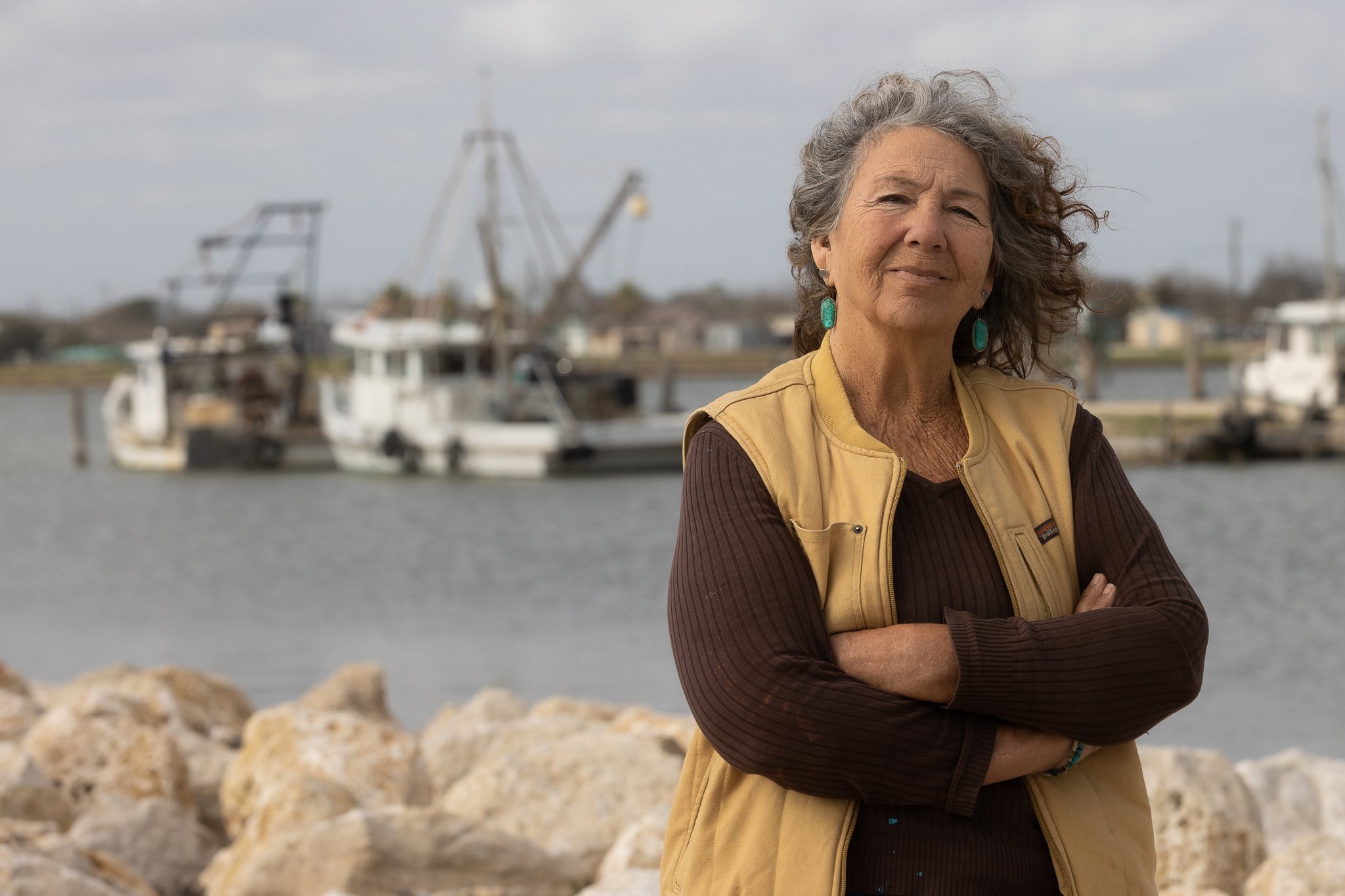 Diane Wilson, a gray-haired woman, stands on a rocky coast on the Gulf of Mexico with shrimp boats in the background.