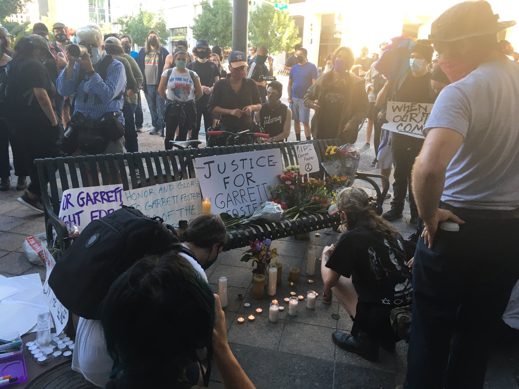 Dozens of people gather around a park bench on an Austin city street at a corner just by where Garrett Foster was murdered by Daniel Perry. Signs line the park bench saying Justice for Garrett Foster, while candles burn underneath.