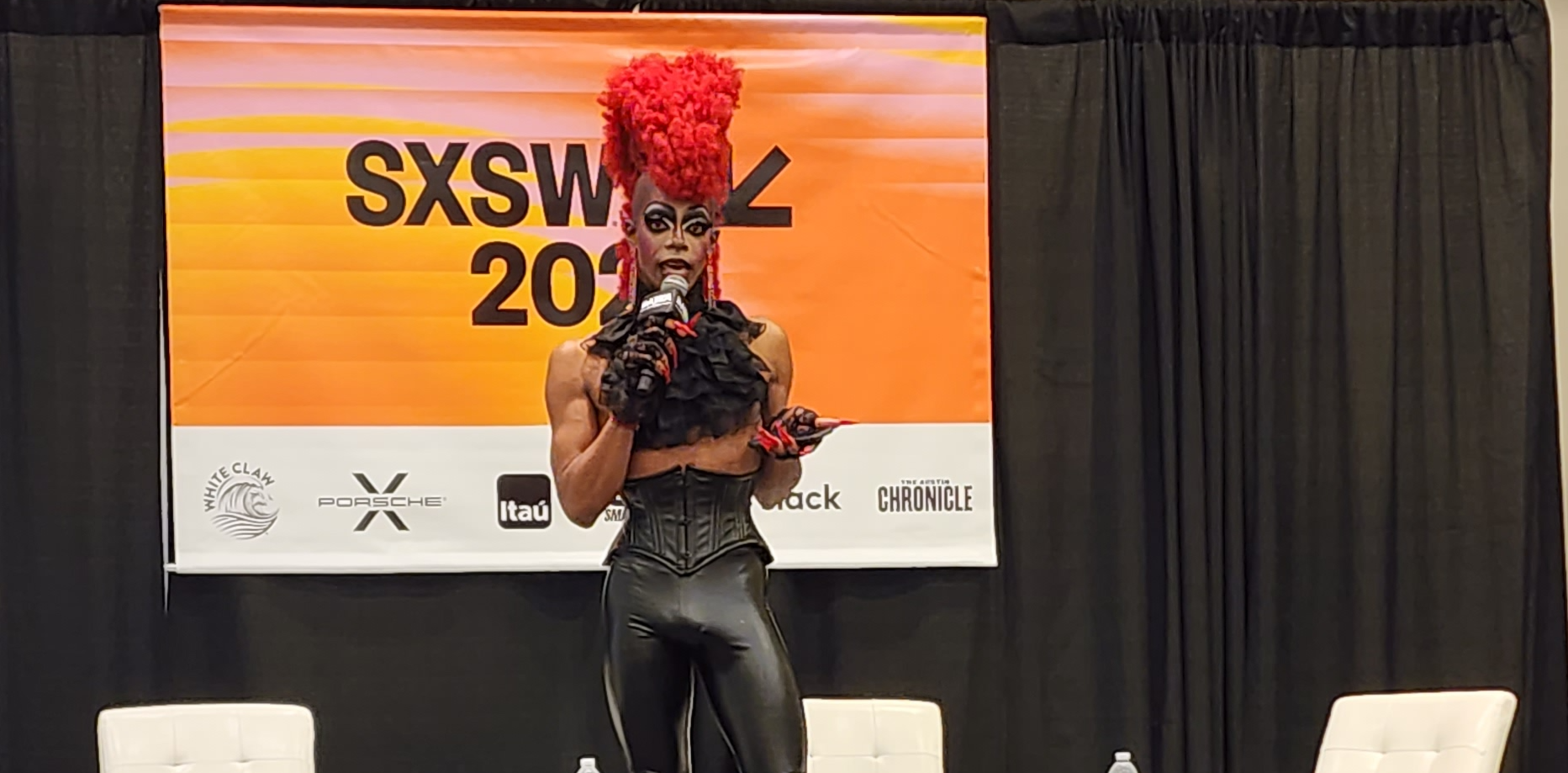 A Black drag performer in a flamboyant red wig, tight leather leather pants, and corsetted top stands in front of a SXSW 2023 banner.