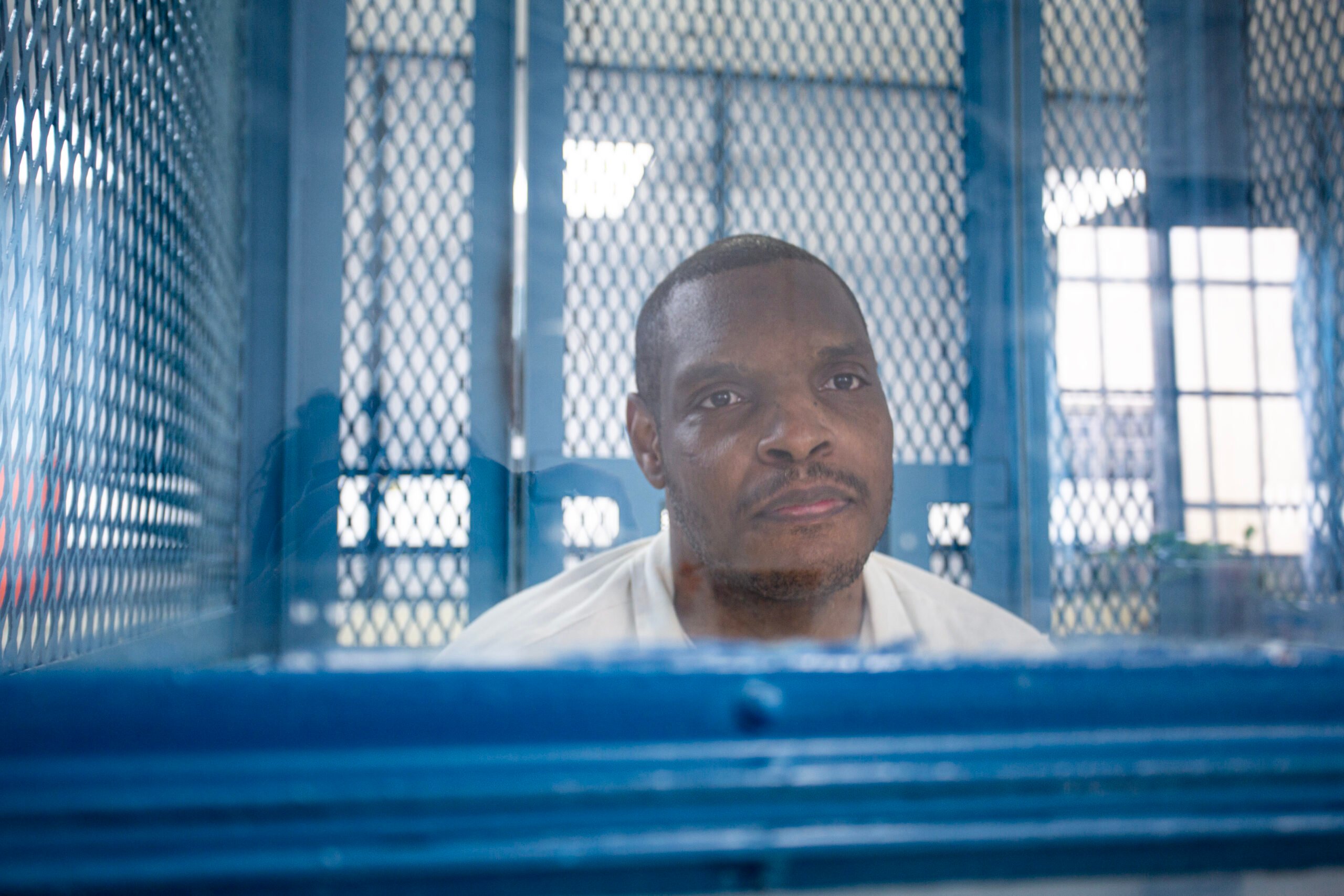 Prison journalist Jason Walker, a Black man with short hair and a short mustache. He has a serious expression on his face and is wearing a collared prison shirt, sitting behind a visitation window in this photograph.