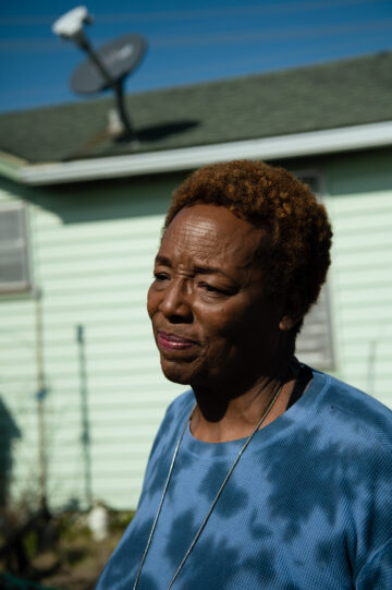Pam Tilly, a Black woman in a all dark blue, tye-dye style sweatshirt frowns pensively, seen in closeup. She's standing in front of the mint green siding and satellite dish of a small home.