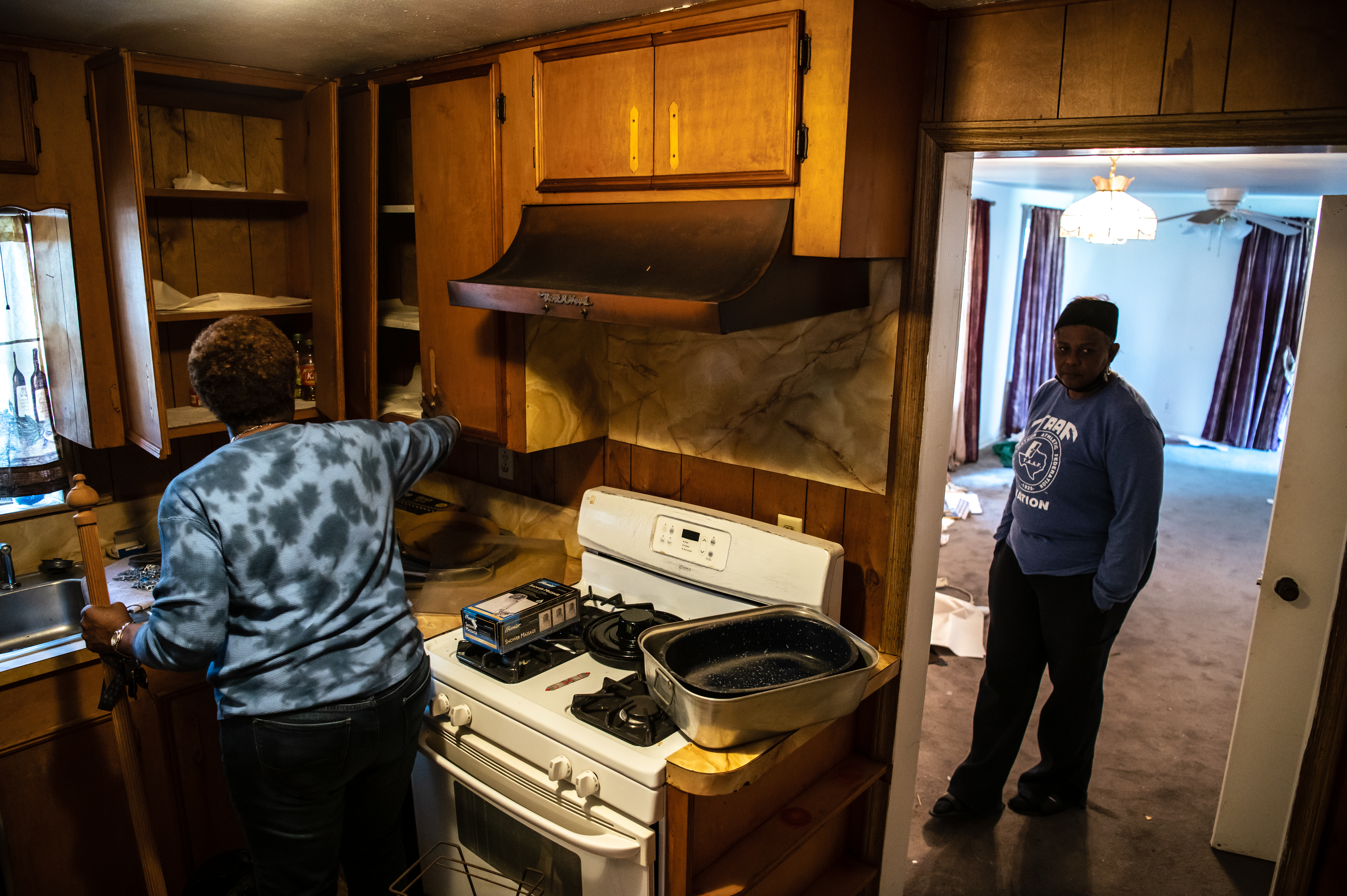 Pam Tilly, a Black woman, has her back to the camera as she packs up the cabinets in an outdated looking kitchen. She has a large pepper mill in one hand. Another family member looks on from the doorway of an empty carpeted room, already packed.