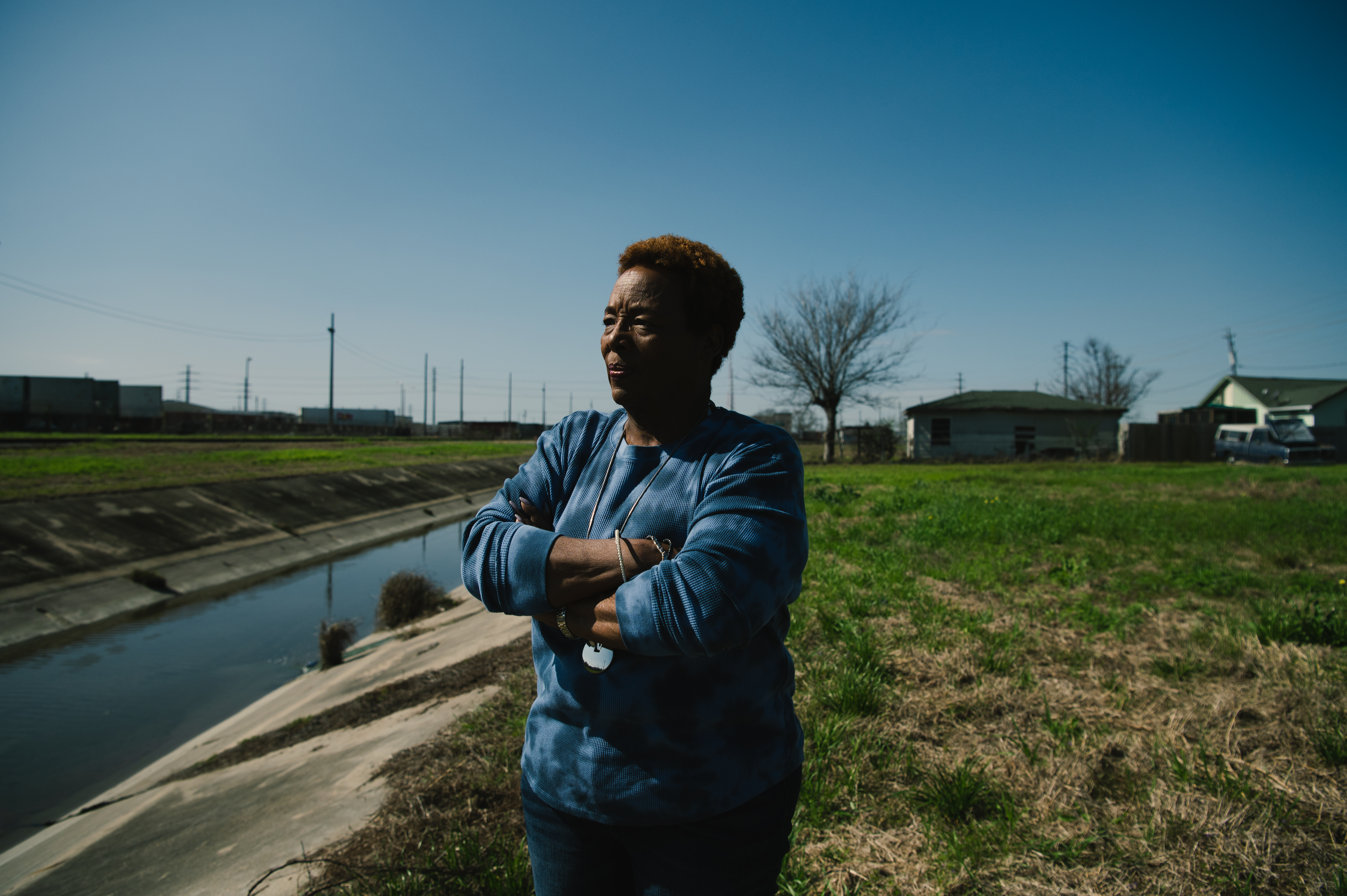 Pam Tilly, a Black woman stands on the bank of a small river in Freeport, Texas with a grassy field and a neighborhood behind her, homes which will soon be lost to the Port of Freeport.