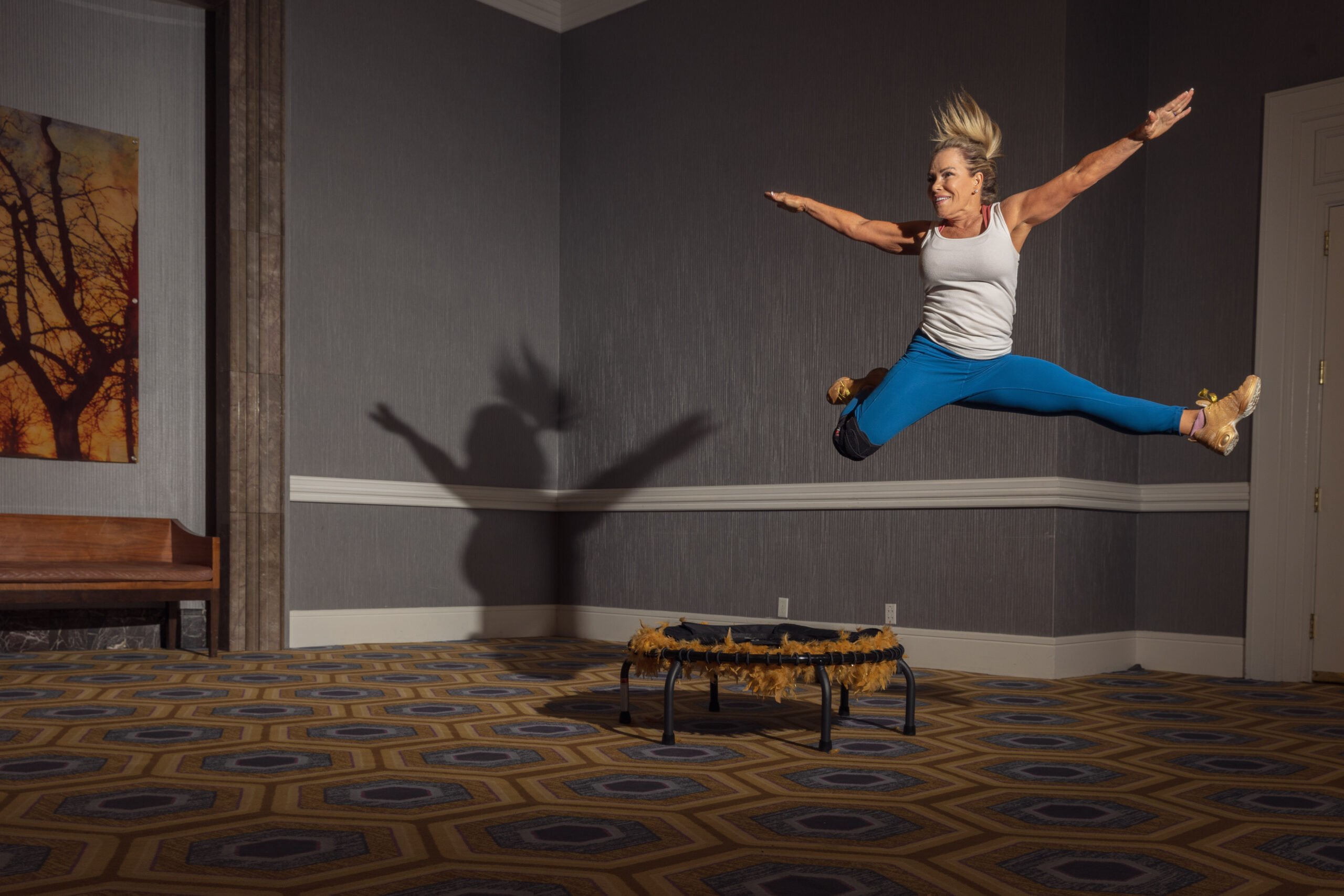 A blonde-haired older woman leaps with arms and legs extended from a trampoline, wearing simple workout gear. She's in a fancy looking hallway.
