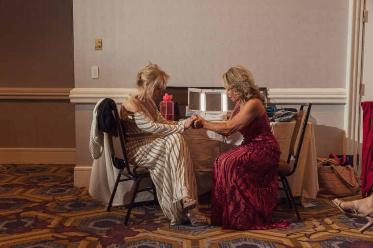 Two blonde older women in evening wear join hands as they pray, seated together.