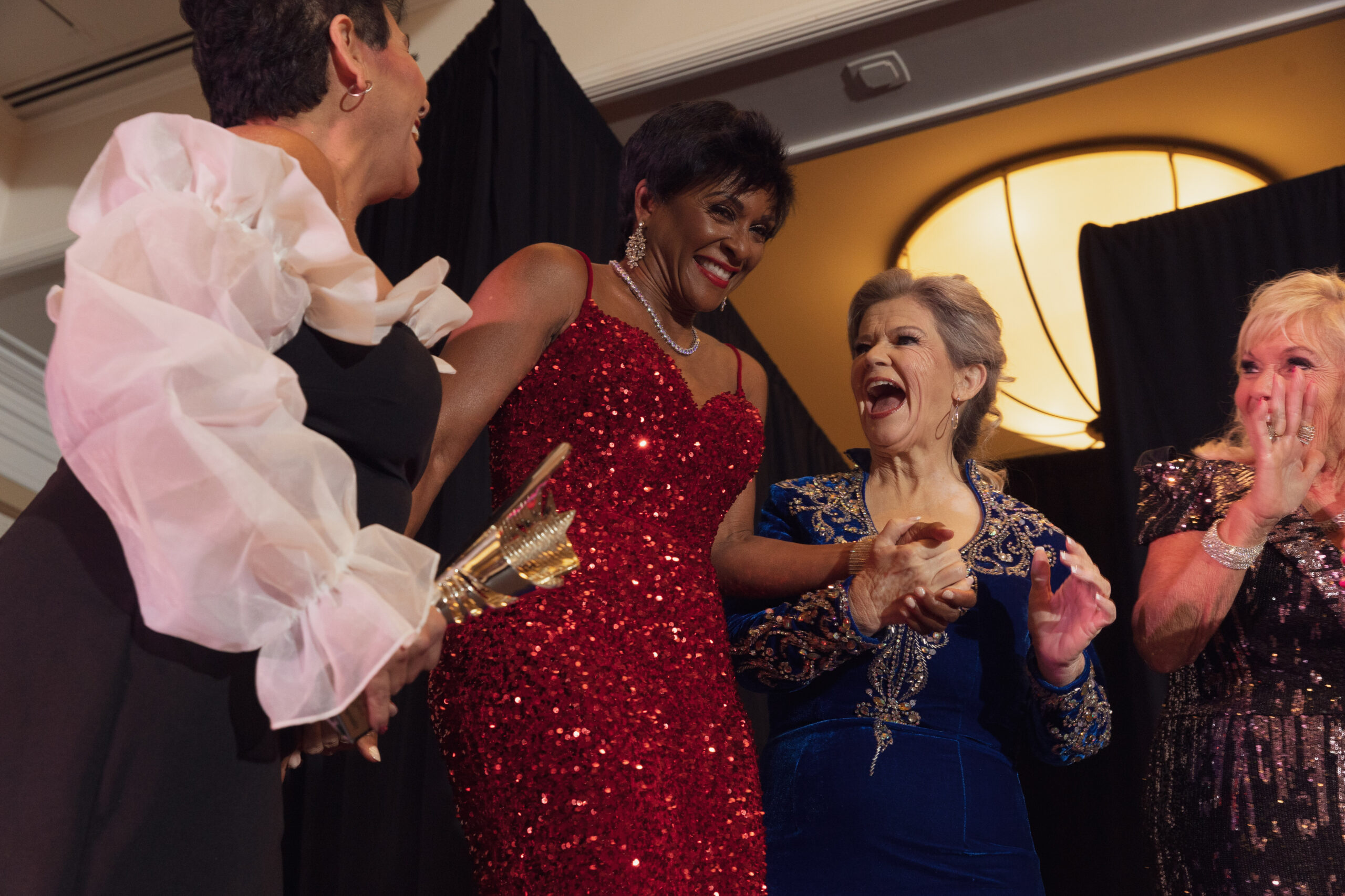A Black woman in a red sequined dress grins as three other woman laugh and celebrate her victory as Ms. Texas Senior America alongside her.
