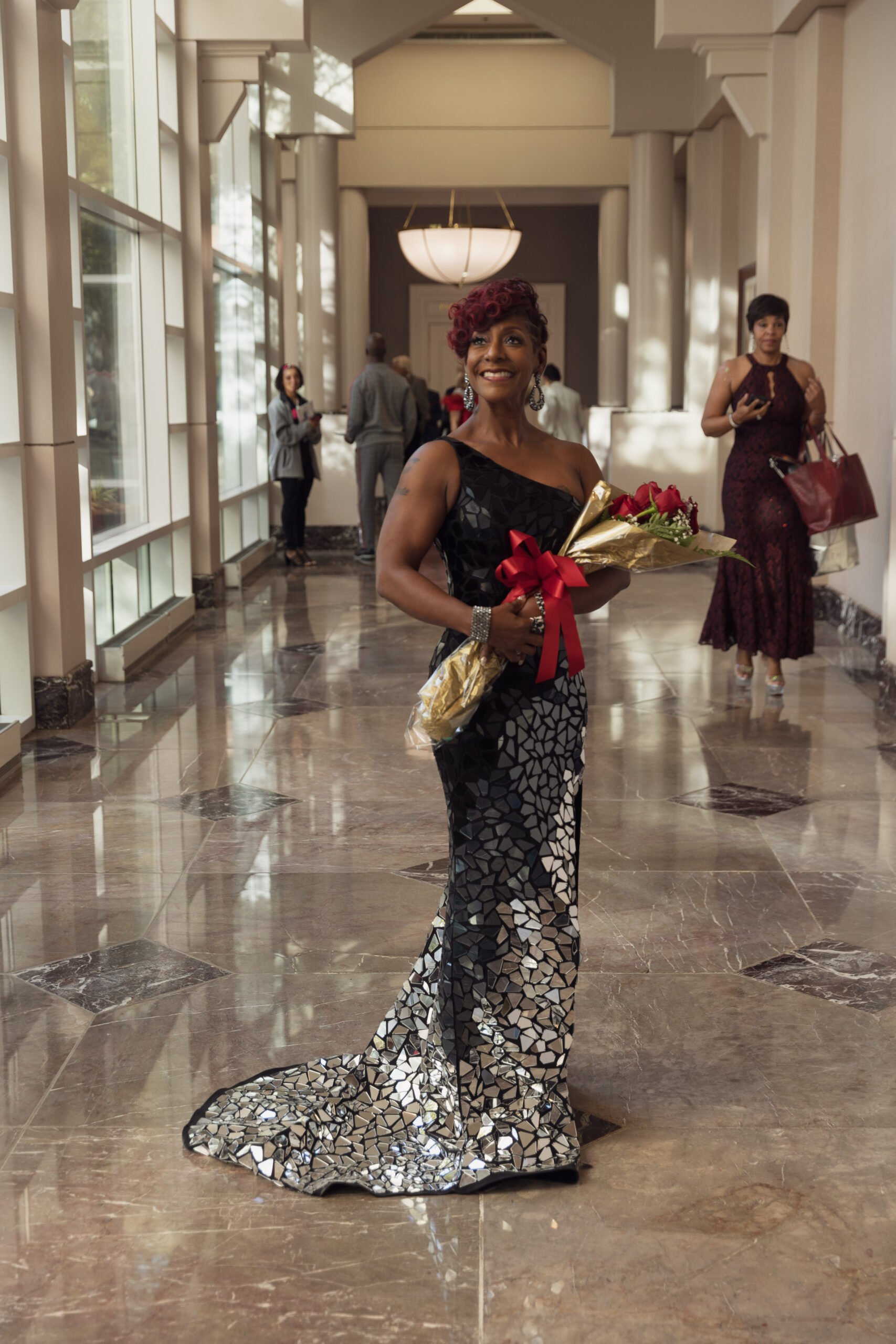 A Black woman in an elegant black gown with a silver mirrored mosaic around its skirts, holds a bunch of roses in her hands as she beams with pride, standing in an elegant tiled hallway.