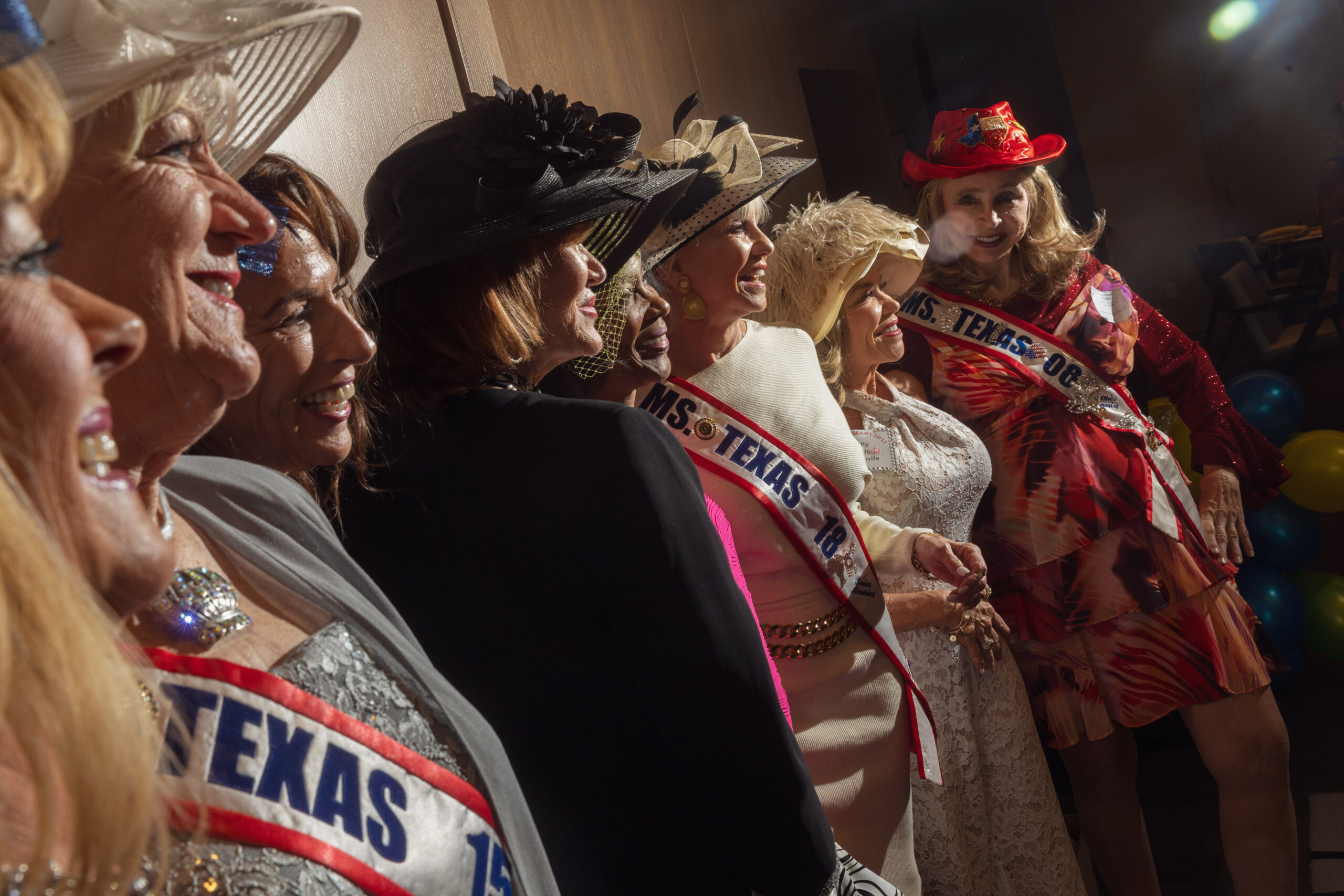 A row of older women in formal evening gowns and sashes announcing the years they won the Ms Texas Senior America pageant.