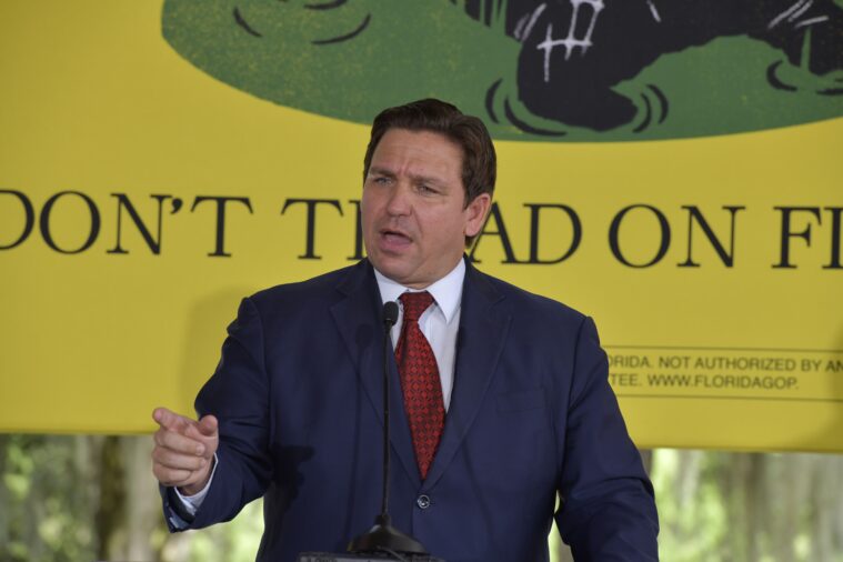 Wearing a suit and tie, Ron DeSantis gestures dramatically while making a political speech. He's standing in front of a banner that has modified Gadsden flag imagery, reading "Don't Tread on Florida."