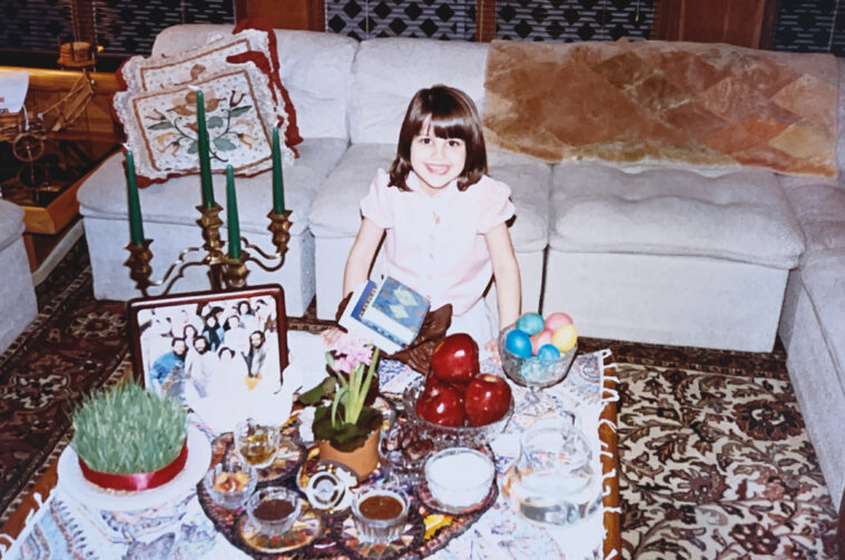 A young girl smiles, seated on a carpet in front of a sumptuous Iranian feast, candles, and other celebratory items.