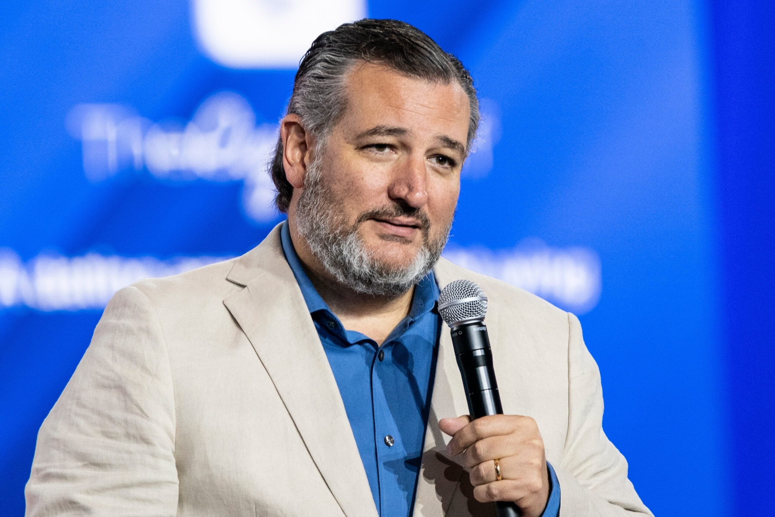 Ted Cruz, wearing a light suit jacket over a blue button-down, speaks into a microphone at CPAC in Dallas.