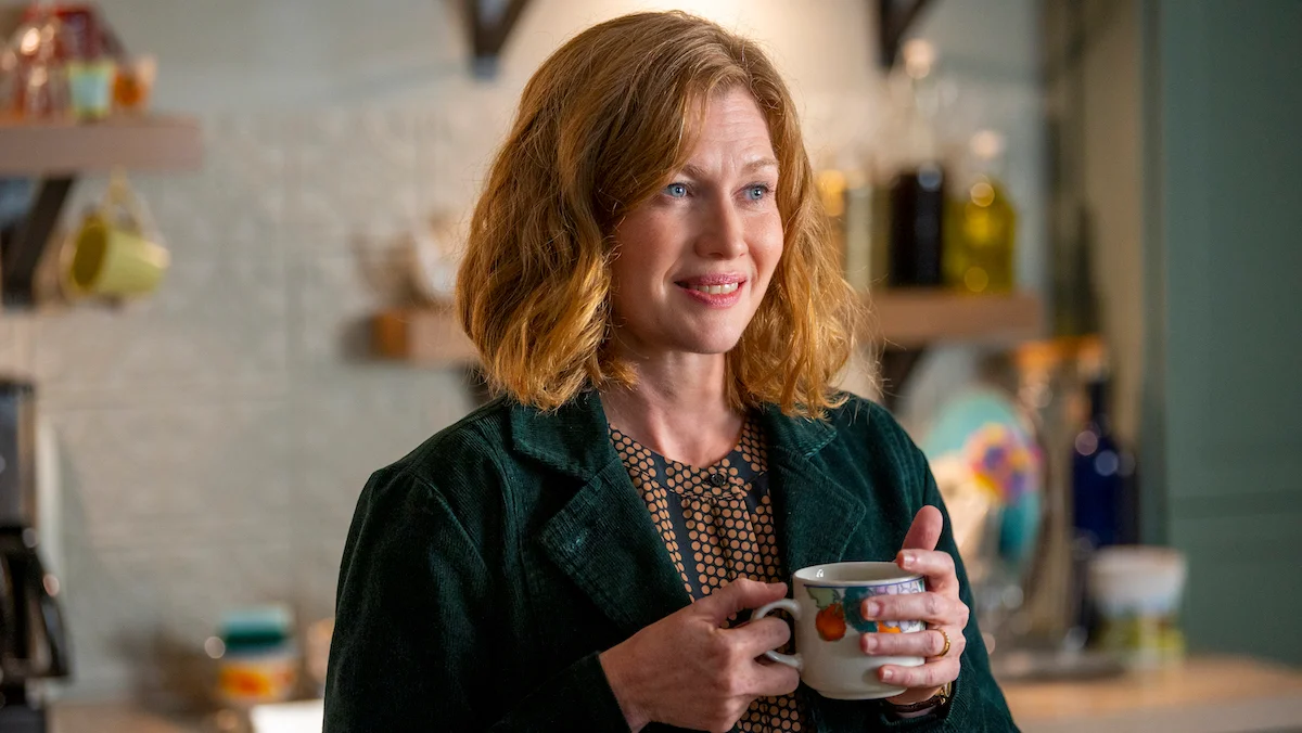 Mireille Enos, a smiling white woman with dirty blonde hair about shoulder length. She's holding a coffee cup in both hands while wearing a dotted blouse under a dark green corduroy jacket.