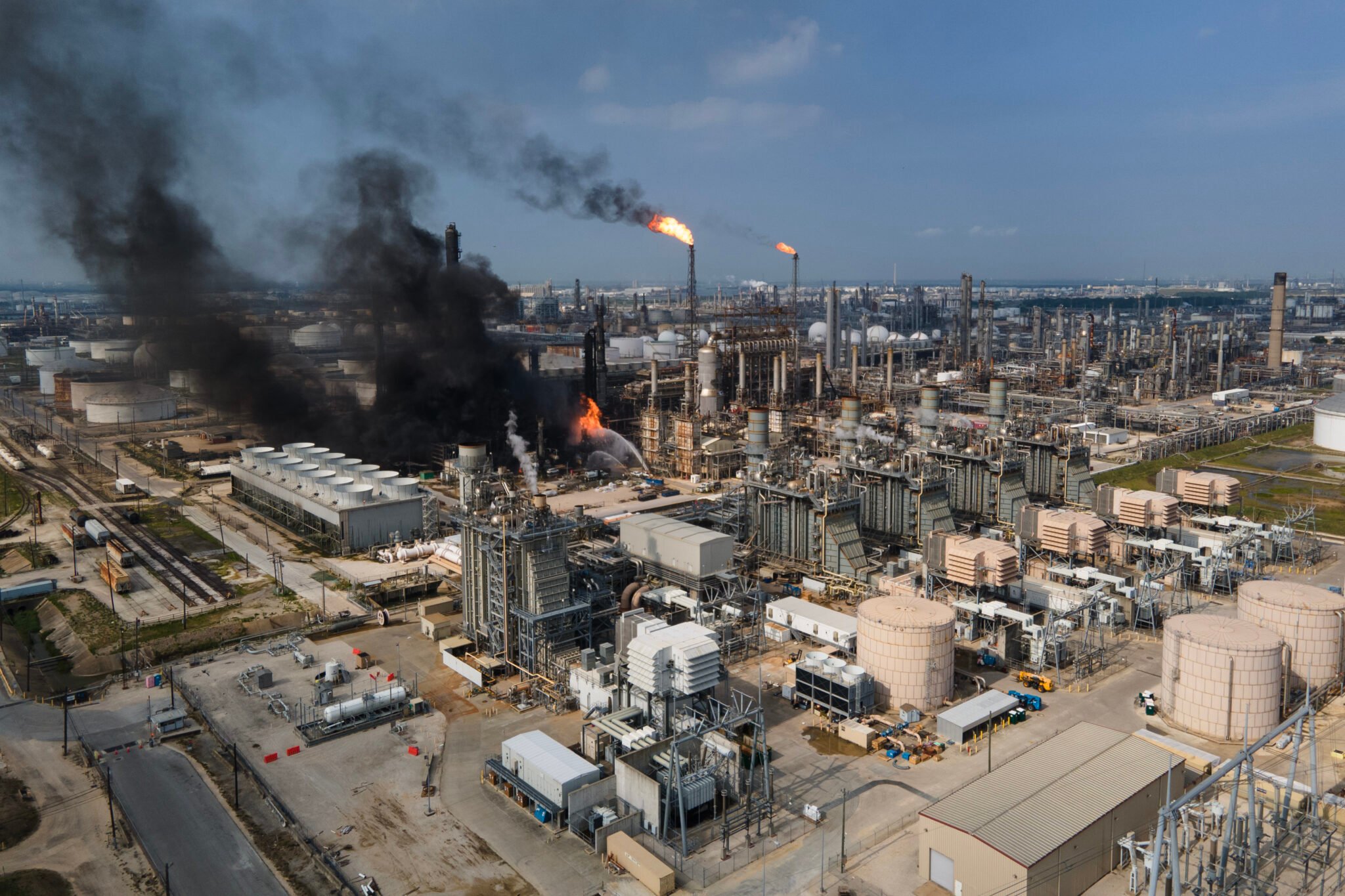 Plumes of black smoke rise into the sky from a massive fire at a Houston-area Shell refinery. Streams of water from firefighters are also visible while smokestacks burn off other chemicals.