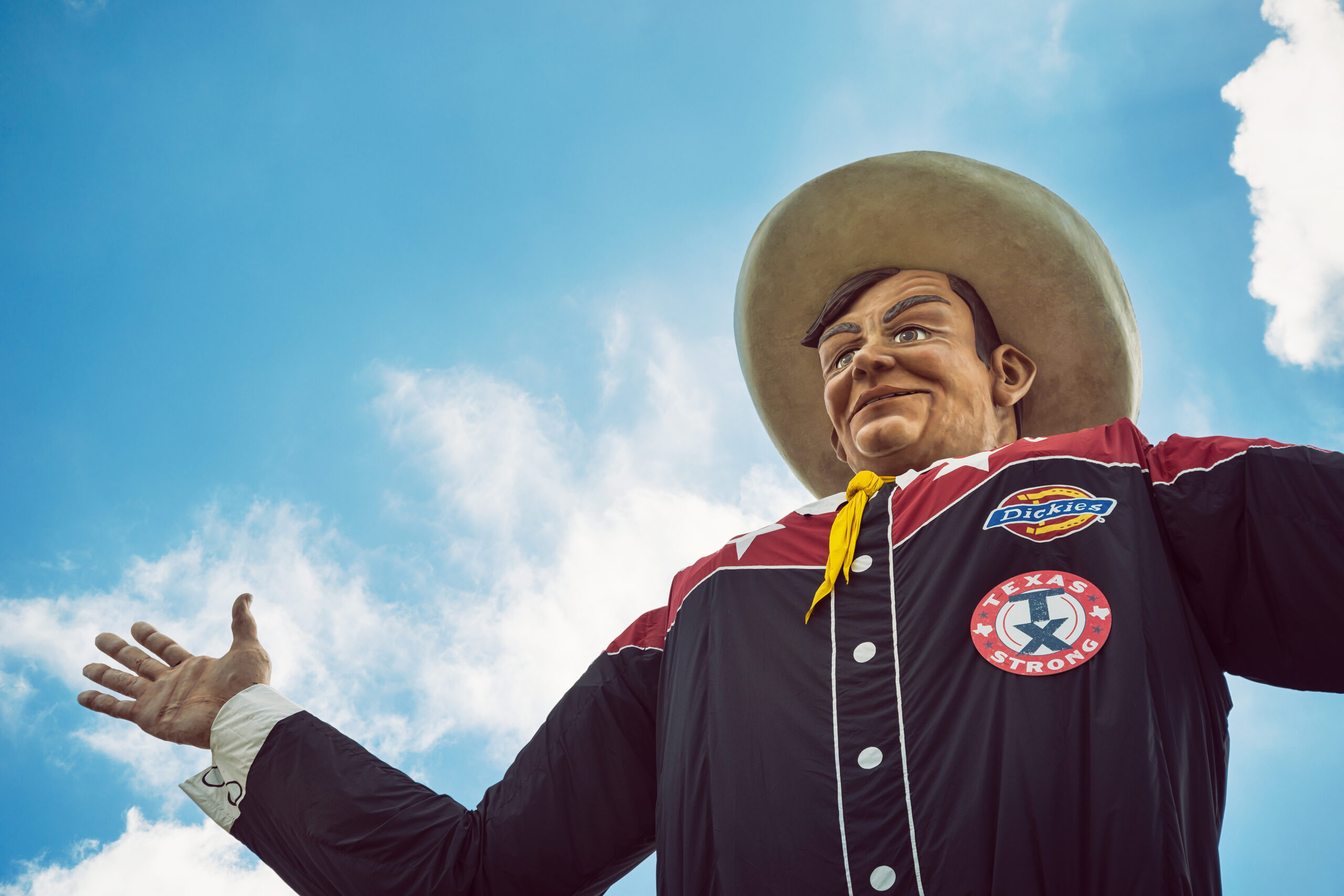The torso, face and arm of the giant Big Tex statue at the Texas State Fair. The cowboy statue is seen from beneath, under a partly cloudy blue sky.