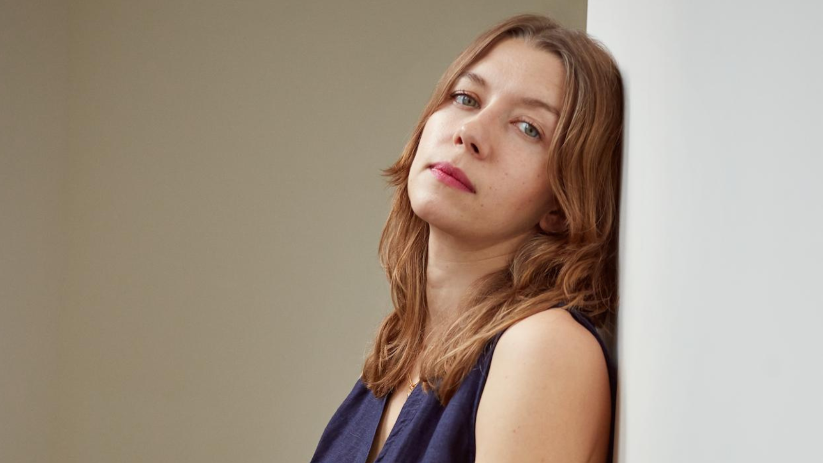 Szilvia Molnar, a blonde woman in a blue dress, leans against a wall while looking at the camera.