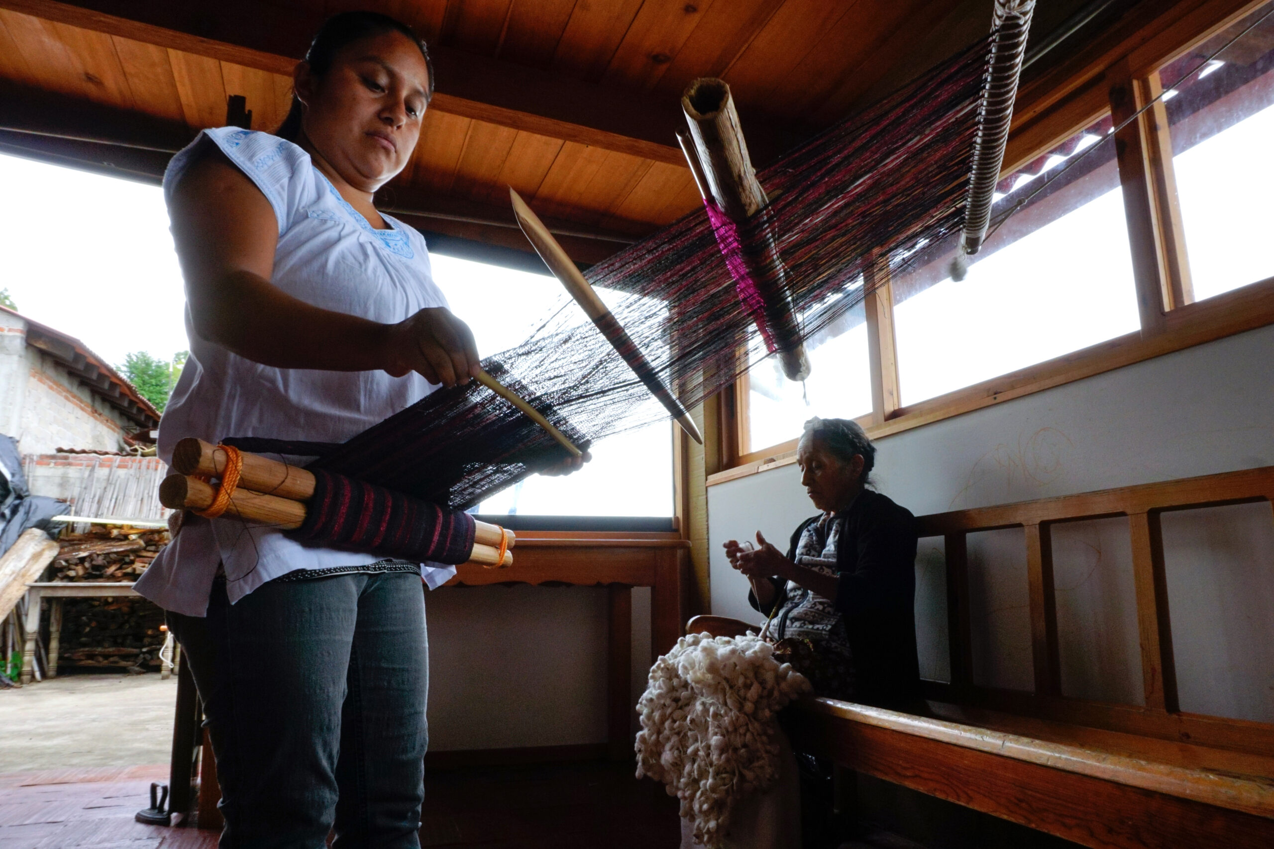 Daysi GarcÌa, left, weaves silk on a backstrap loom while her grandmother, Margarita Flores, spins silk thread at MoisÈs MartÌnezís family workshop in San Pedro Cajonos, Mexico, on Nov. 13, 2022. Their building is a simple wooden one with big windows, and another loom is visible outside.