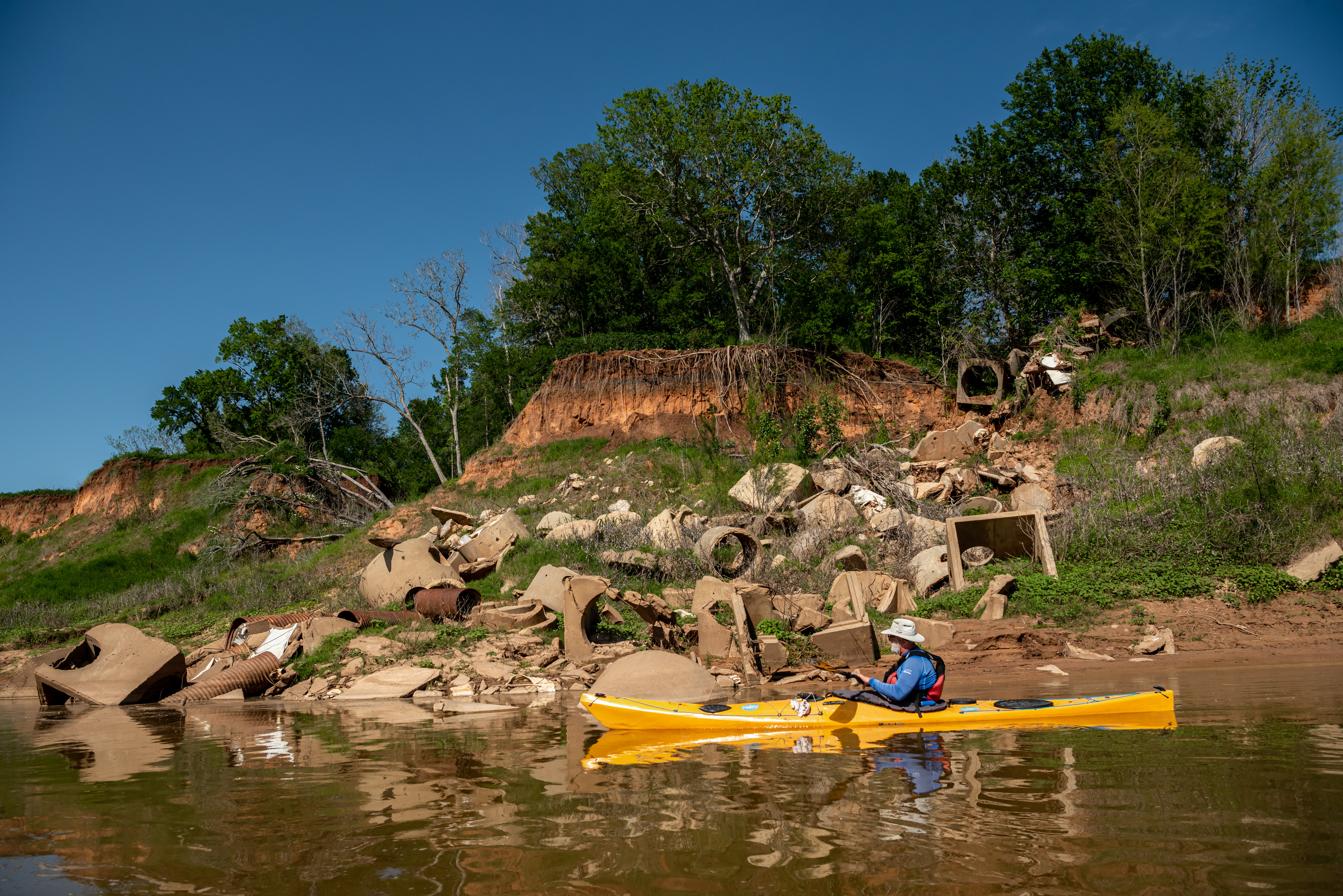 Bruce Bodsn, a man in an outdoor hat and blue parka and red safety vest, navigates a yellow kayak next to a heap of assorted cement refuse lying on a hillside by the Brazos River, under the roots of trees visible along an eroded embankment.
