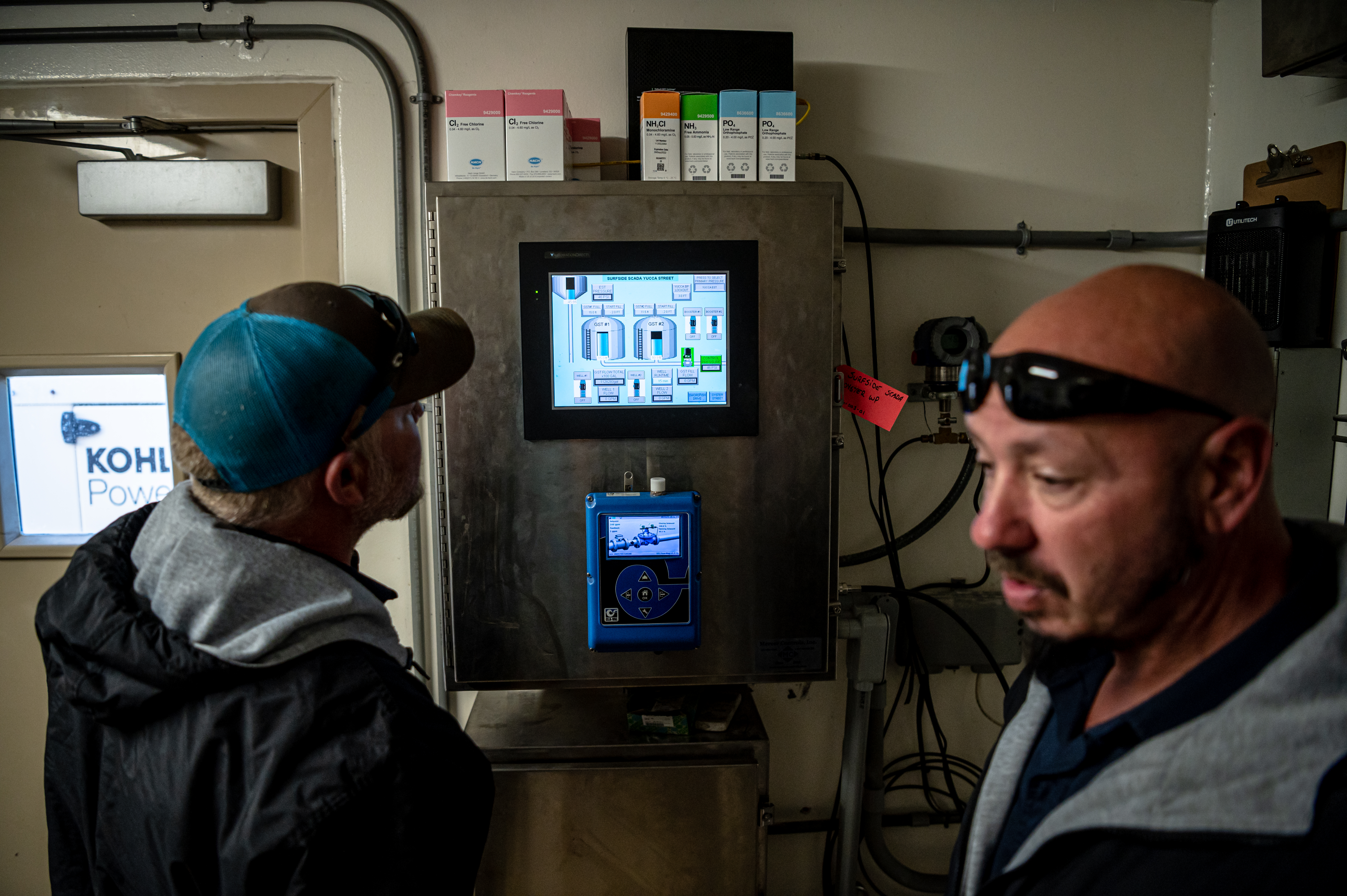 Two men in hoodies, one wearing a ball cap, examine a digital readout on a water pumping system. 