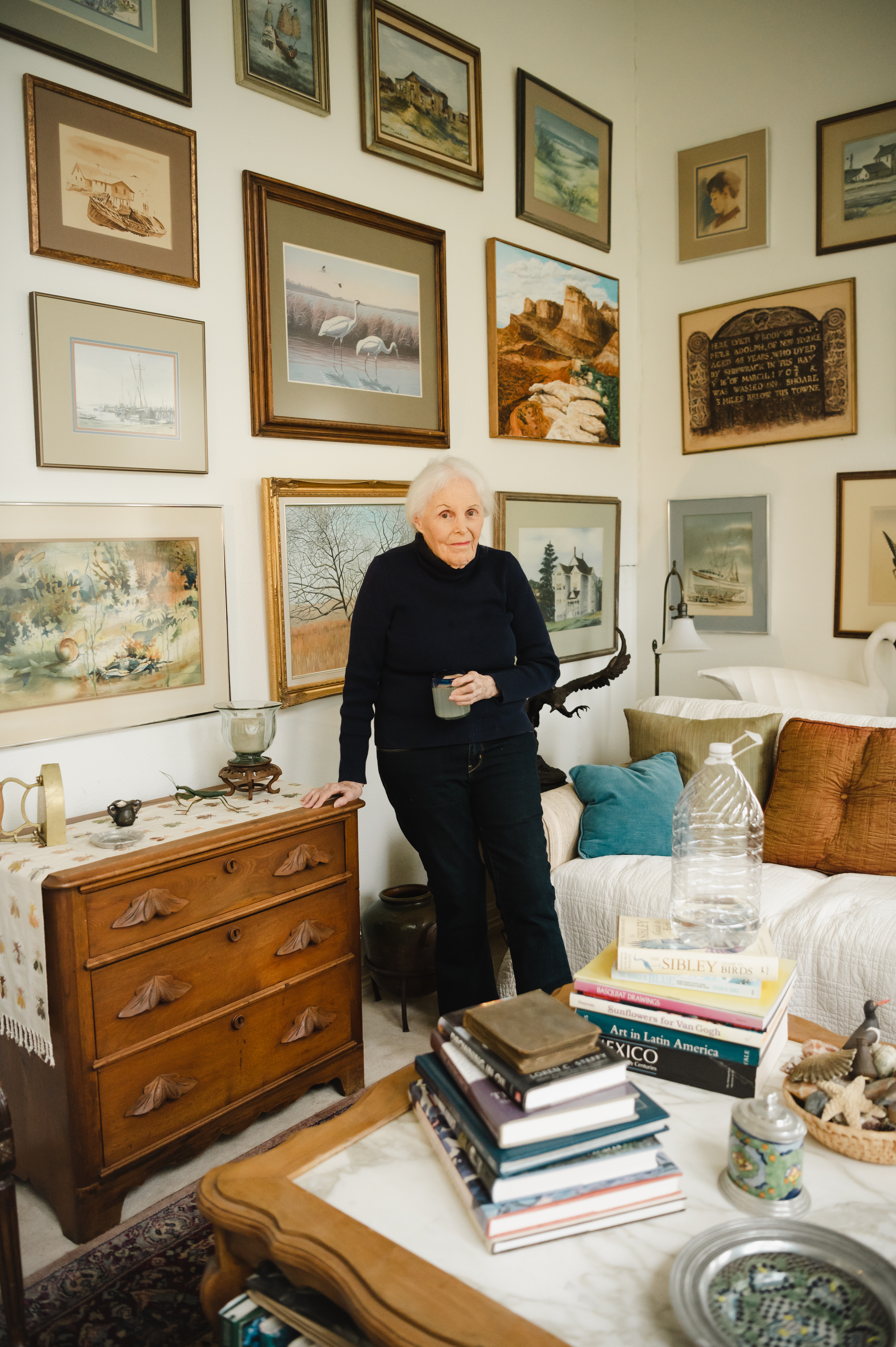 A white haired, elderly white woman poses with a mug in one hand, the other leaning on a dresser in a living room area, the wall covered in framed nature photographs. It's a cozy space, with books and pillows.