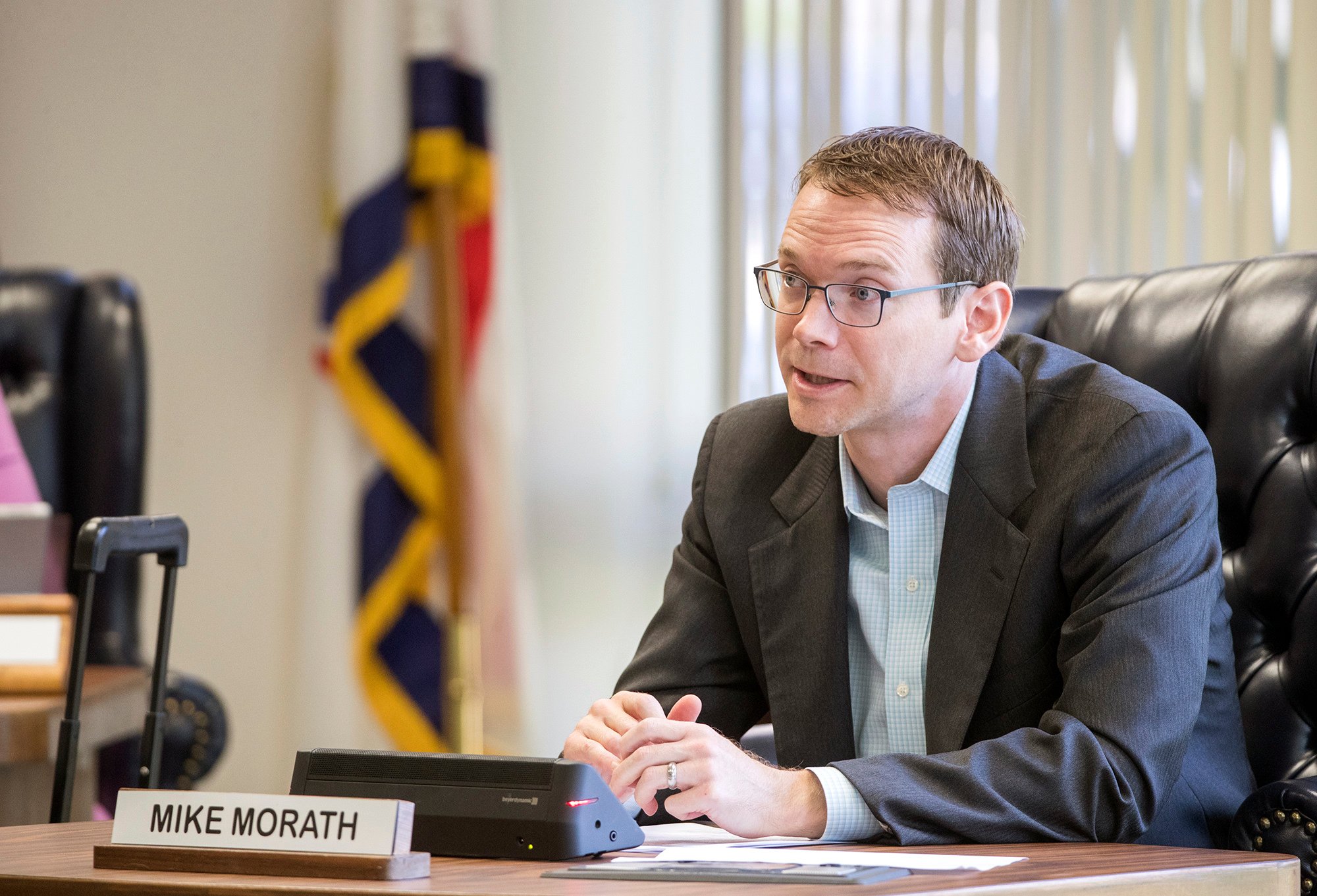 Mike Morath sits in his office, wearing a suit jacket and button-down shirt without a tie. He is a white man with short brown hair and glasses.
