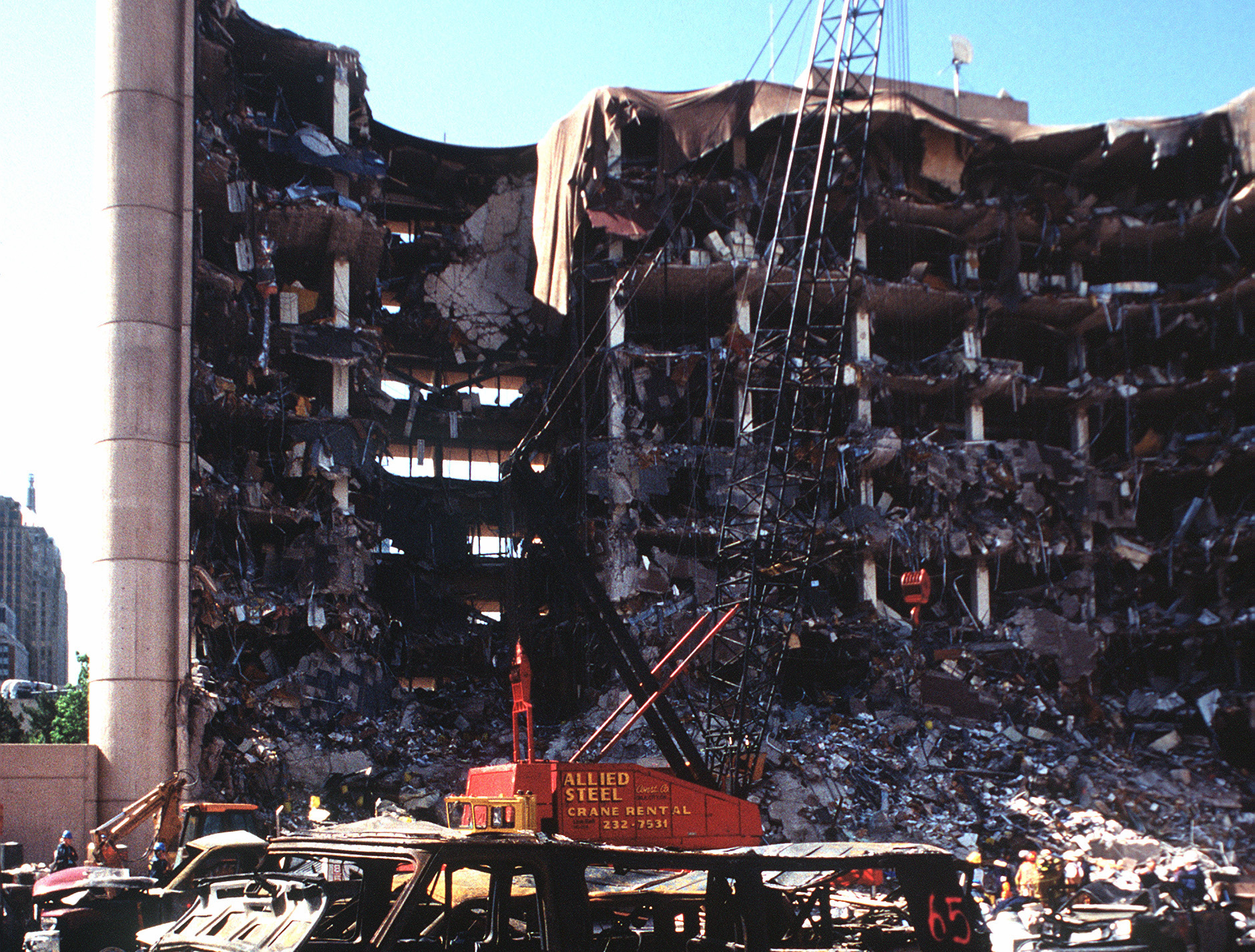 The devastated facade of the Alfred P. Murrah Federal Building in Oklahoma City after Timothy McVeigh's bombing. A burnt out van and a red rescue crane are in the foreground.