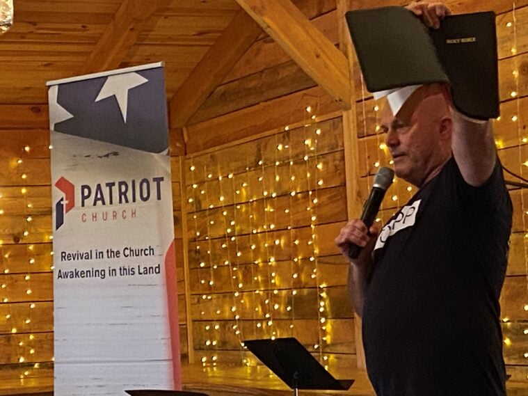 Ken Peters of Patriot Church, a bald white man in a dark shirt, holds a bible dramatically above his head. Nearby is a sign reading "Patriot Church: Revival in the Church, Awakening in this Land."
