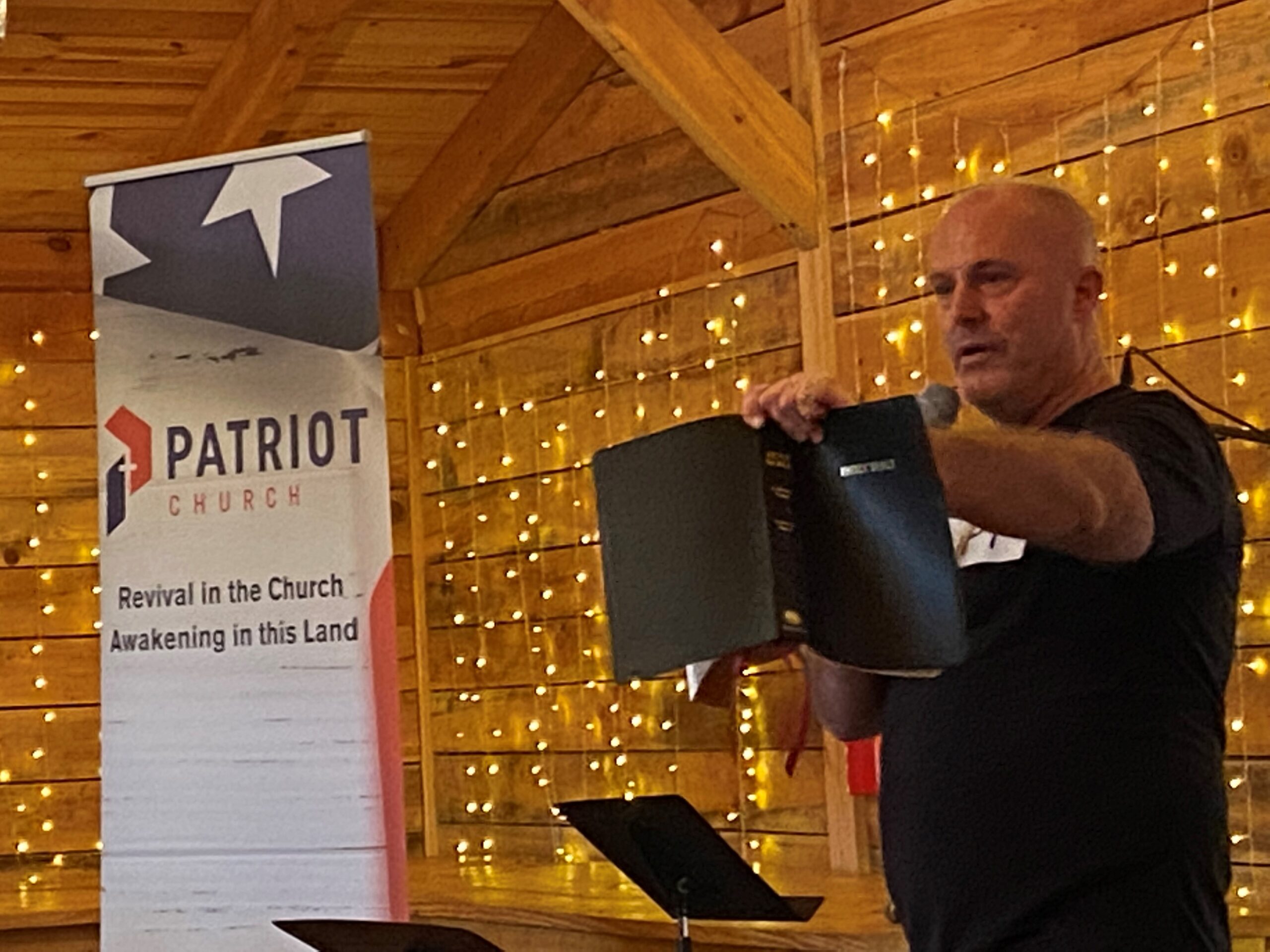 Ken Peters of Patriot Church dramatically holds up a bible near a sign reading "Patriot Church: Revival in the Church, Awakening in this Land."