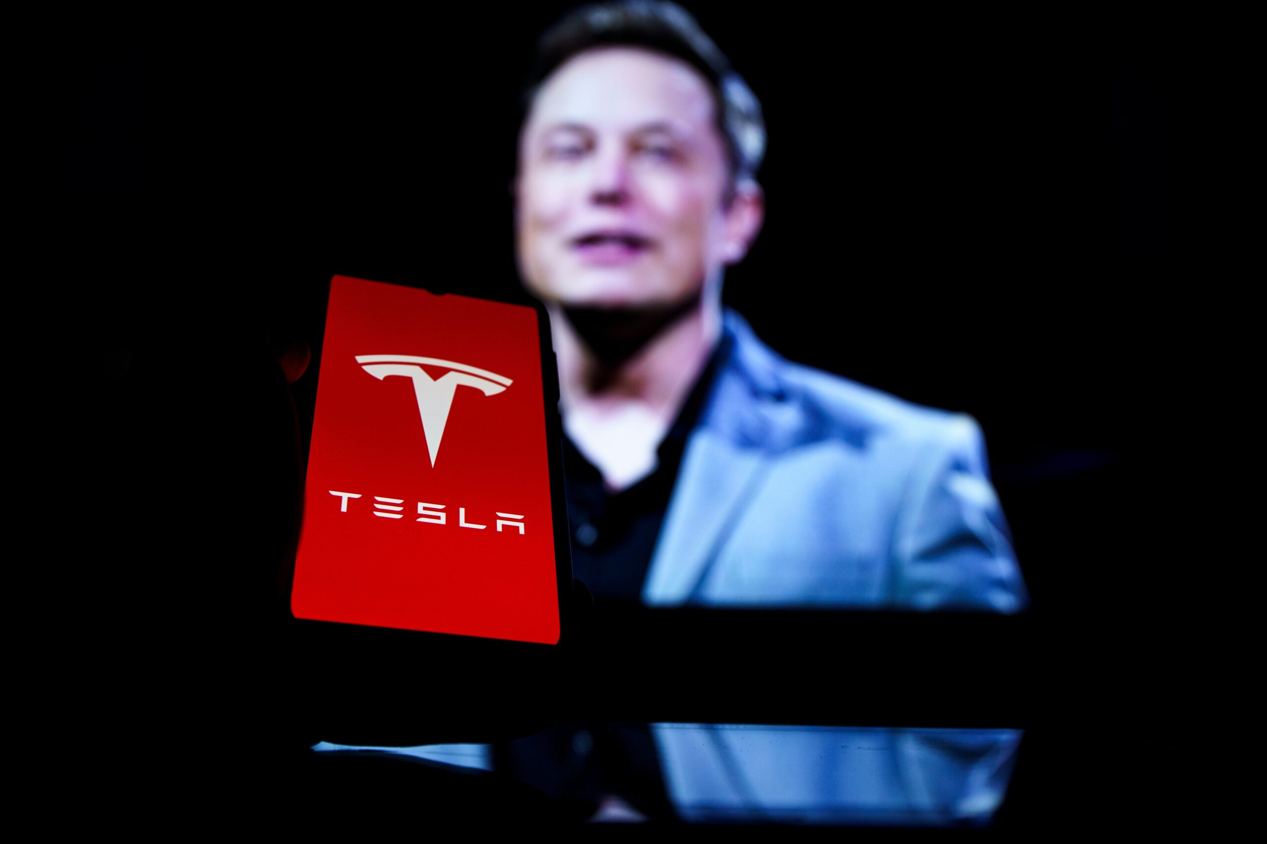 Elon Musk grins behind an electronic device that says "Tesla"
