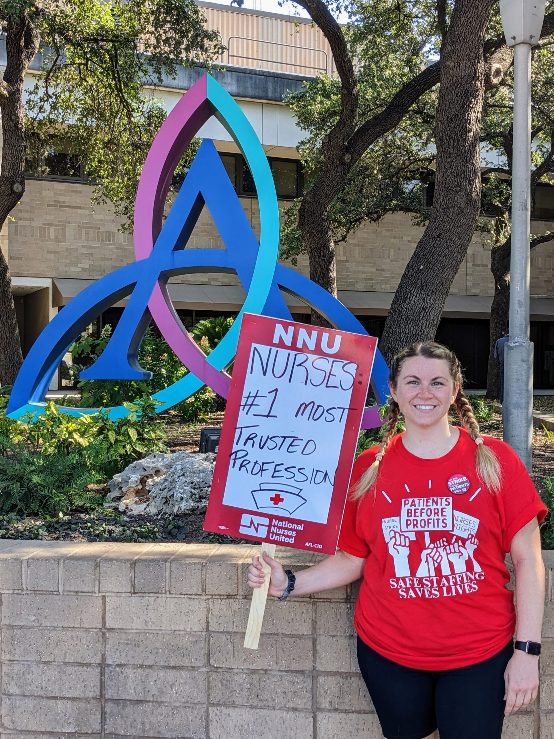 Jessica Gripentrog is a white woman in pig tails, standing in front of the Ascension Seton logo sculpture at the medical center during the nurses strike. She is wearing a National Nurses United red t-shirt and holding a sign that declares nurses to be the number one most trusted profession.