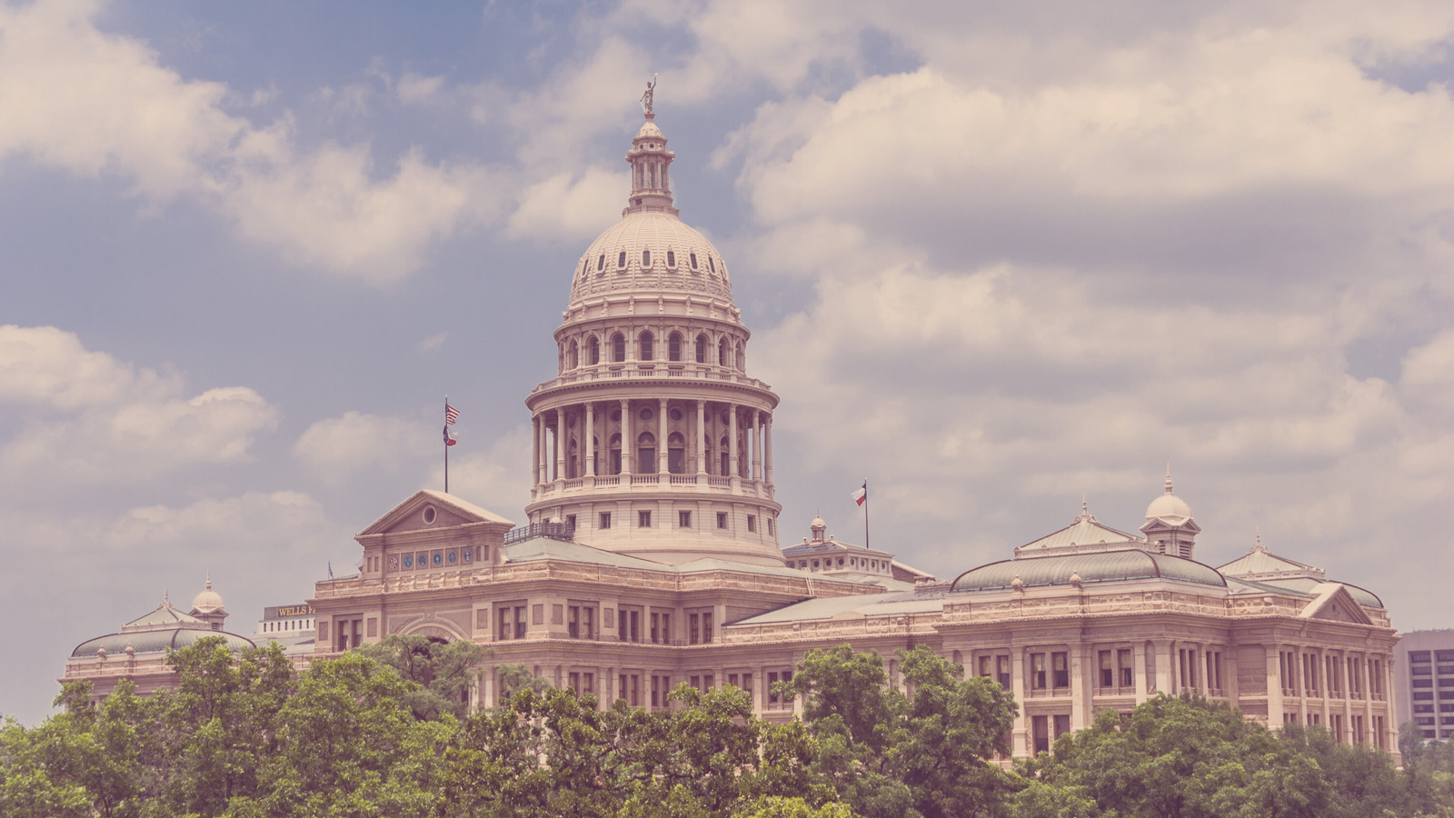 The pink granite dome and main building of the Texas State Capitol, home of the Texas legislature. It's scene under a partly cloudy sky from behind tall green trees. There is a color filter addede to give it a washed out, old timey photograph feel.
