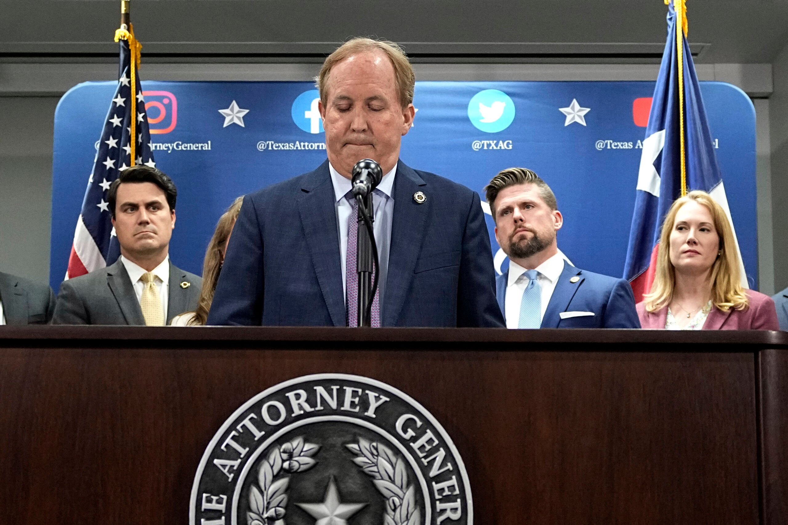 Texas state Attorney General Ken Paxton, center, makes a statement at his office in Austin, Texas. He's wearing a suit and tie as he stands at an Attorney General podium frowning deeply. He's flanked on either side by allies with serious expressions.